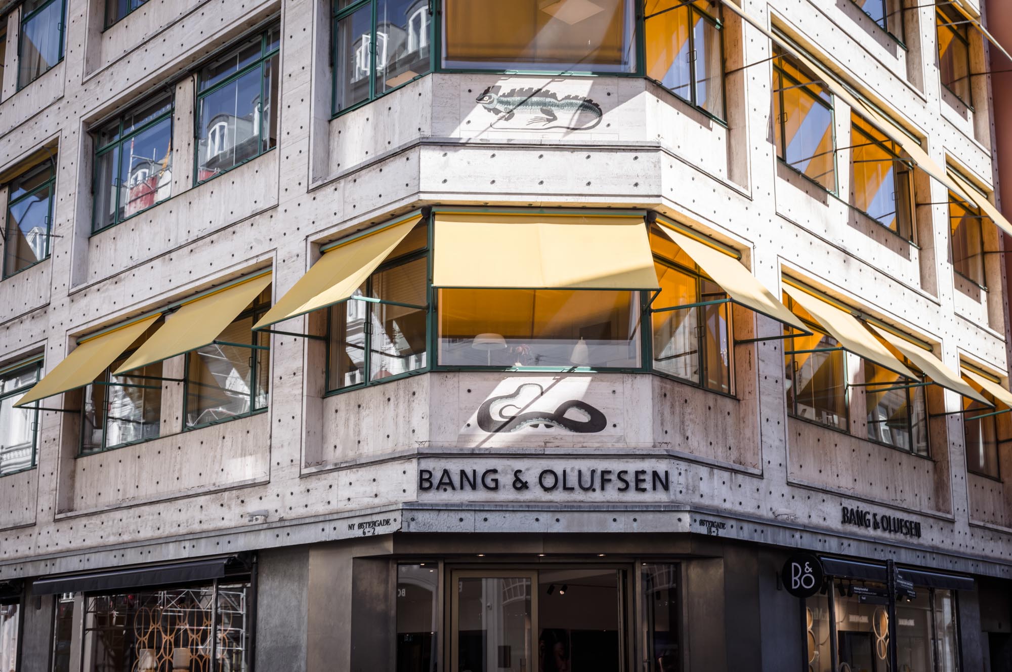 Corner view of a concrete building with yellow awnings and Bang & Olufsen signage, showcasing modern architectural design.