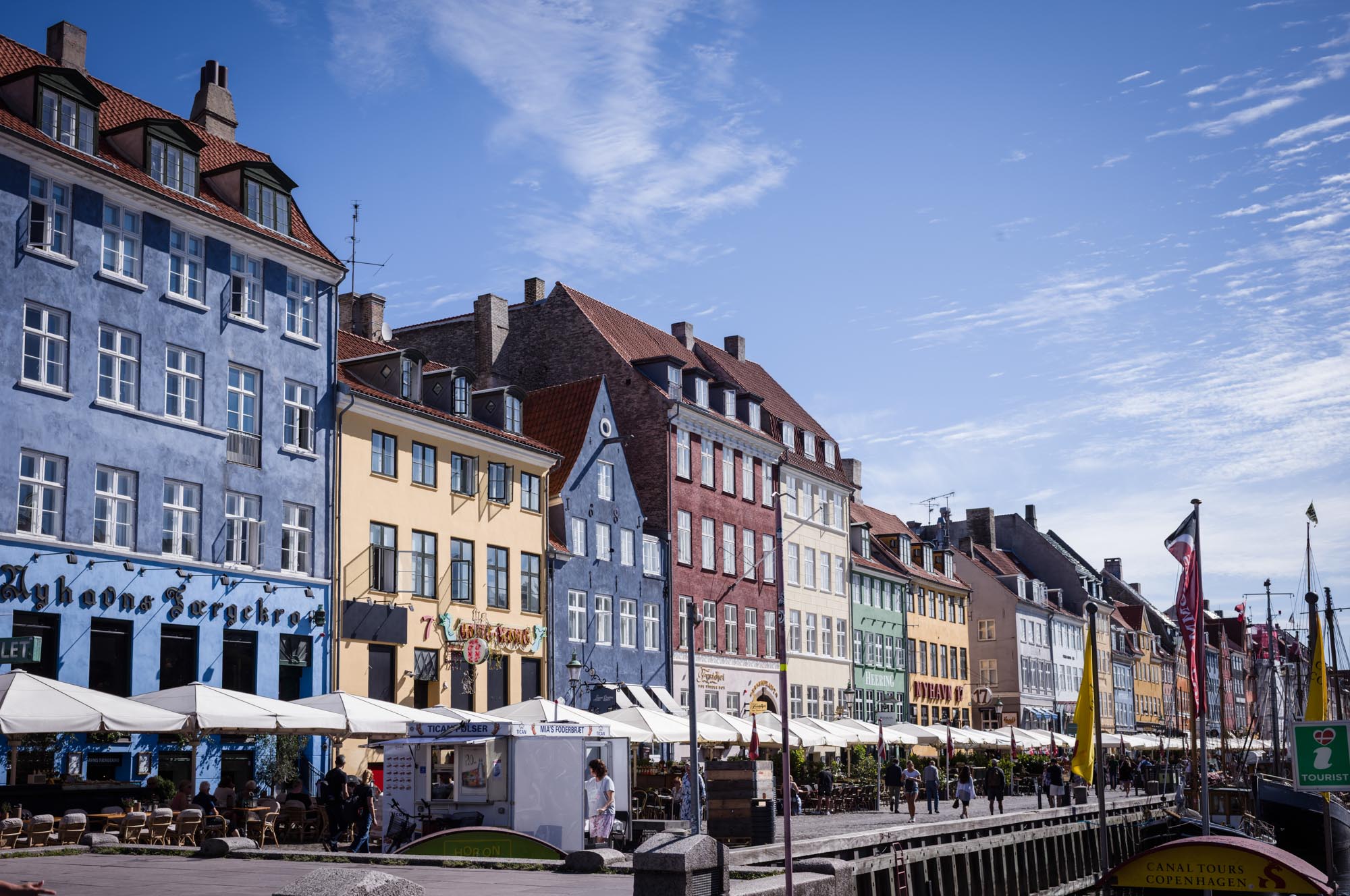 Colorful buildings line Copenhagen's Nyhavn waterfront, bustling with outdoor cafes and visitors under a clear blue sky.