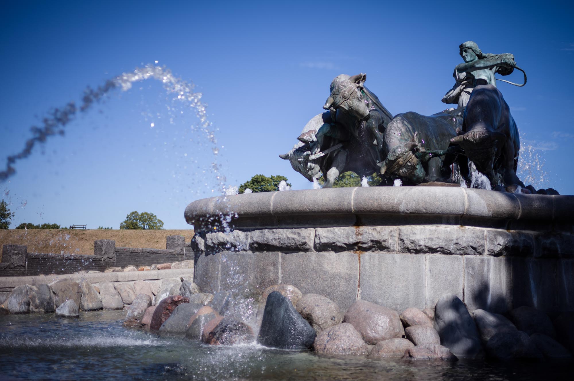 Fountain with bronze sculptures of a woman and oxen, with water jets against a clear blue sky.