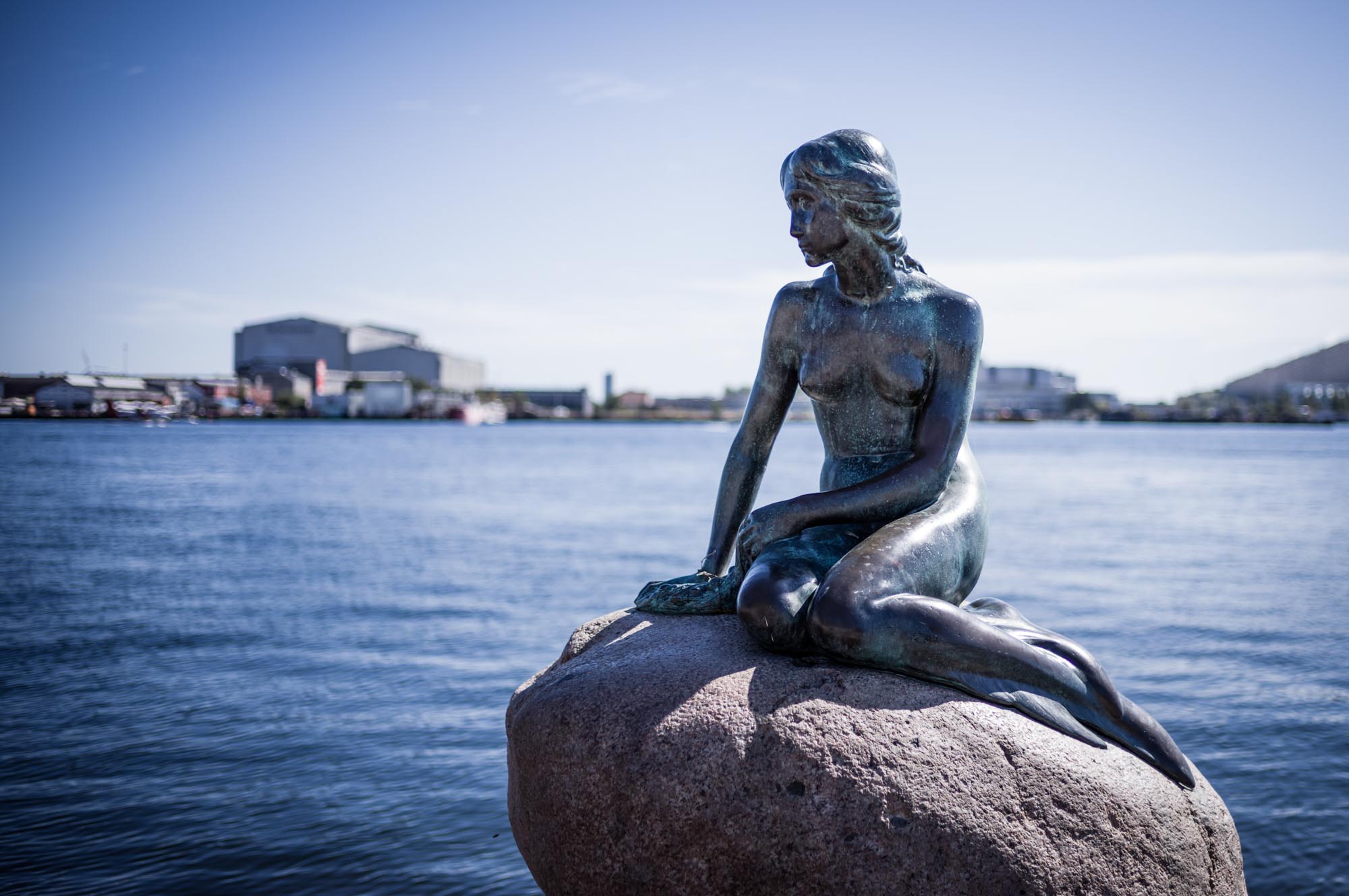 Bronze mermaid statue sitting on a rock near the waterfront with a blurred cityscape in the background.