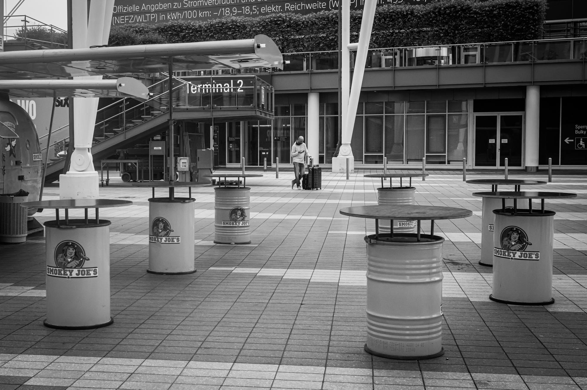 Empty airport terminal with Smokey Joe's tables on a tiled floor, person with suitcase walking in background.