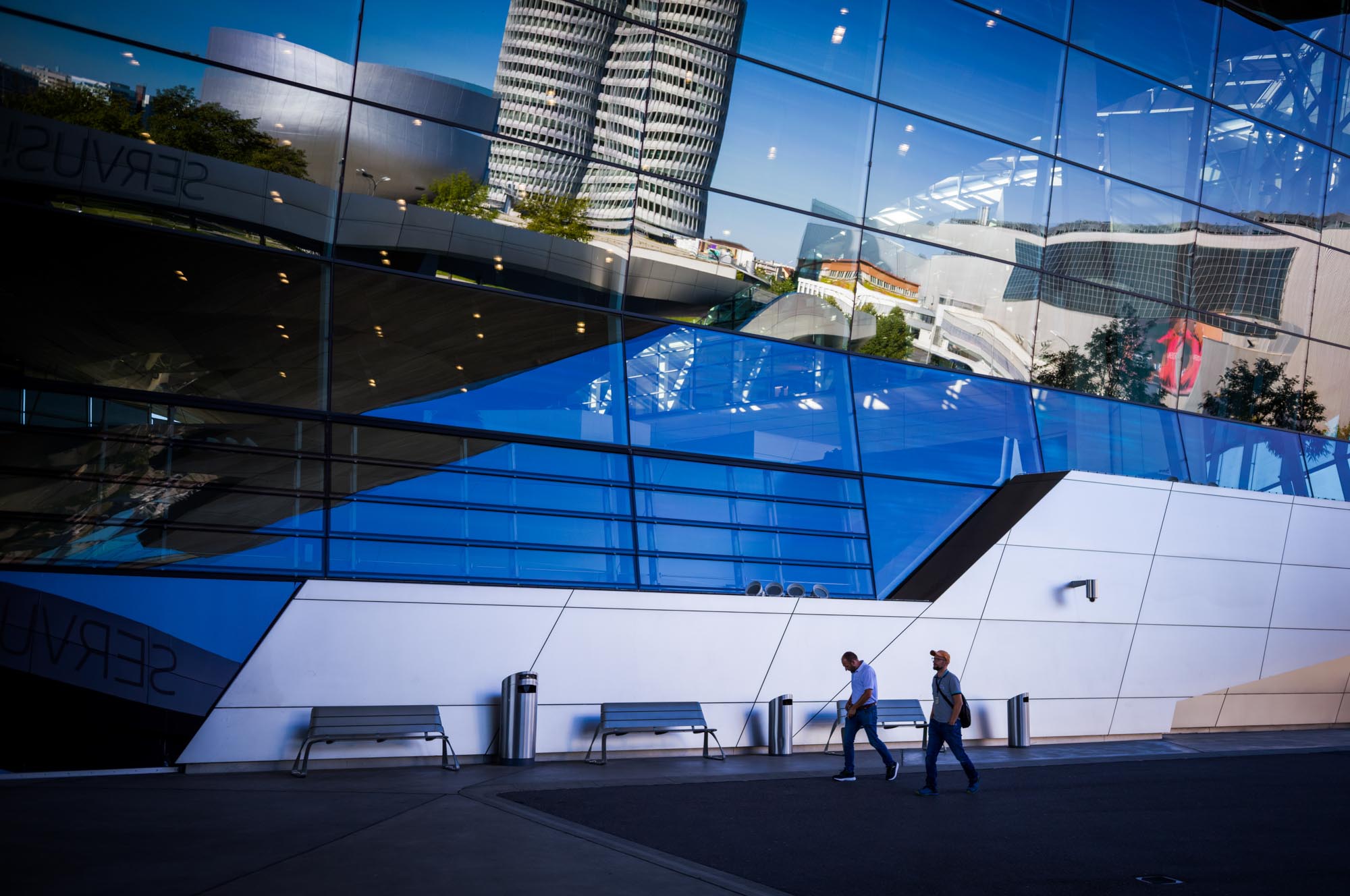 Two people walk past a modern glass building reflecting futuristic architecture in the city.