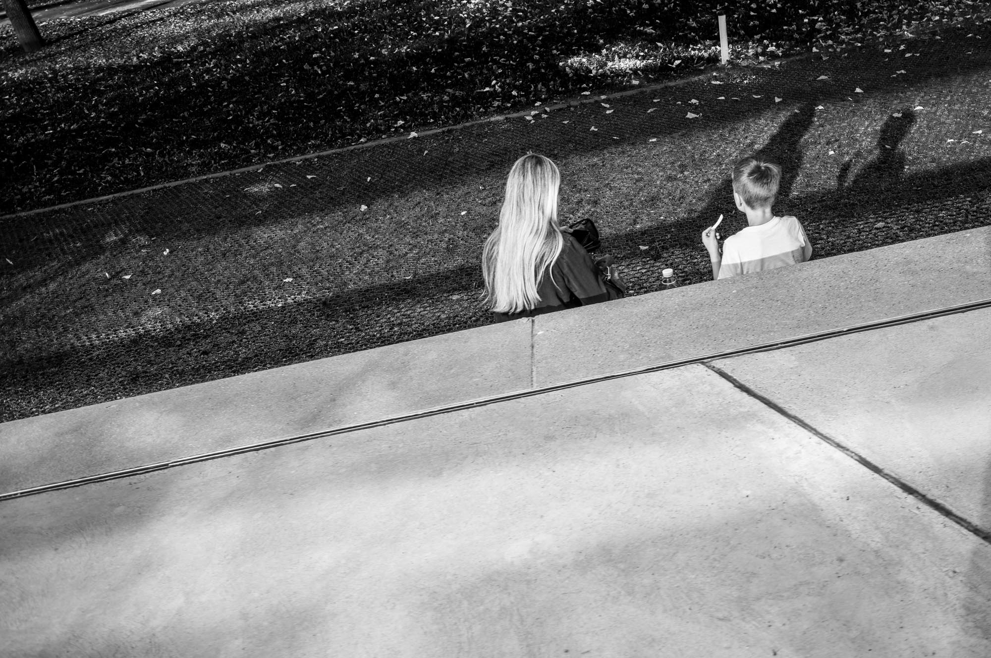 Black and white photo of two people sitting on a bench, casting long shadows on a sunny day.