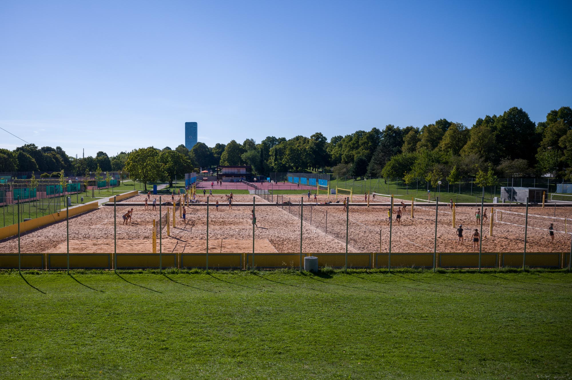 Outdoor beach volleyball courts with players under a clear blue sky, surrounded by greenery and trees.