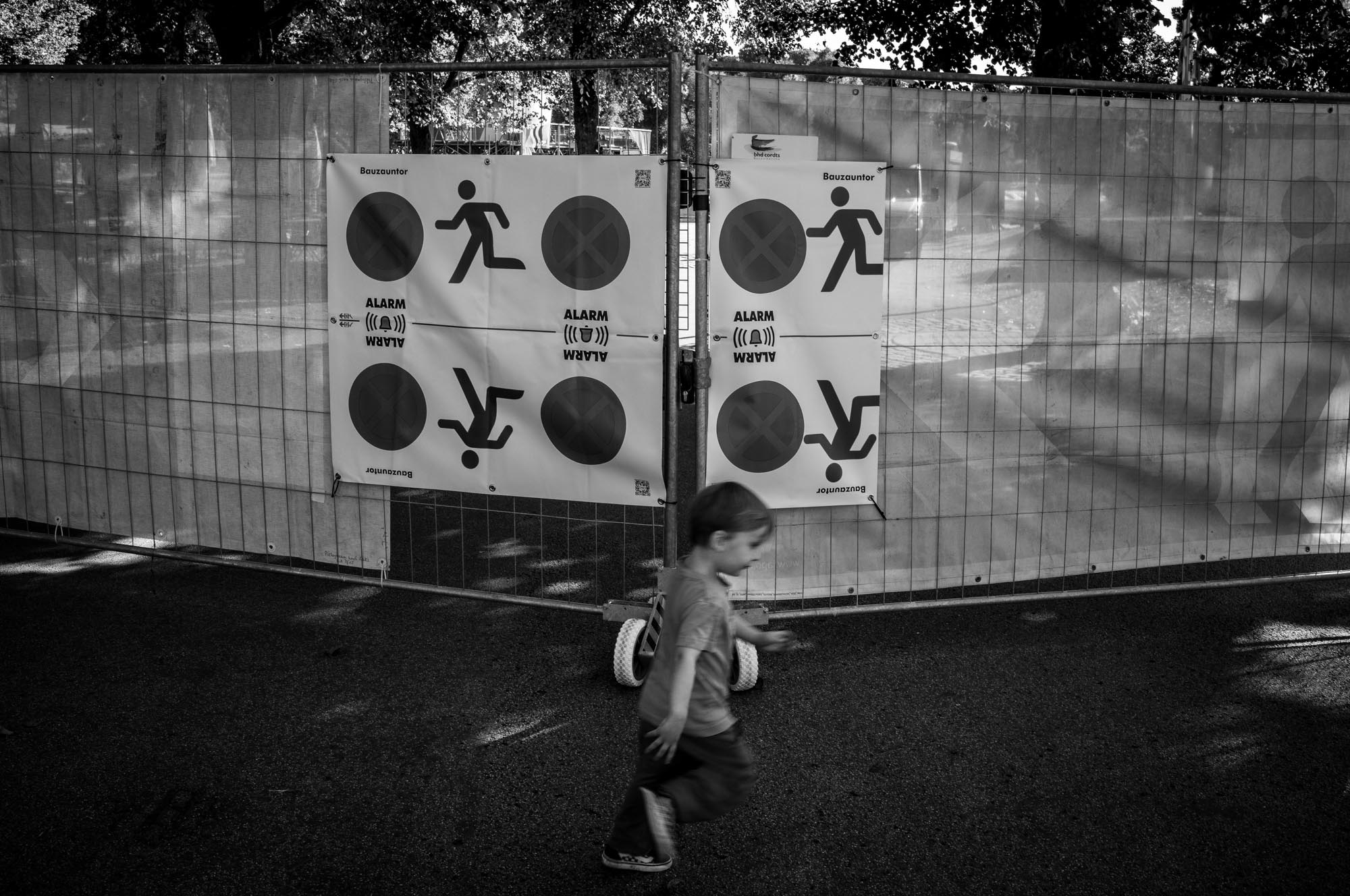 Child walking past construction fence with warning signs in a park.