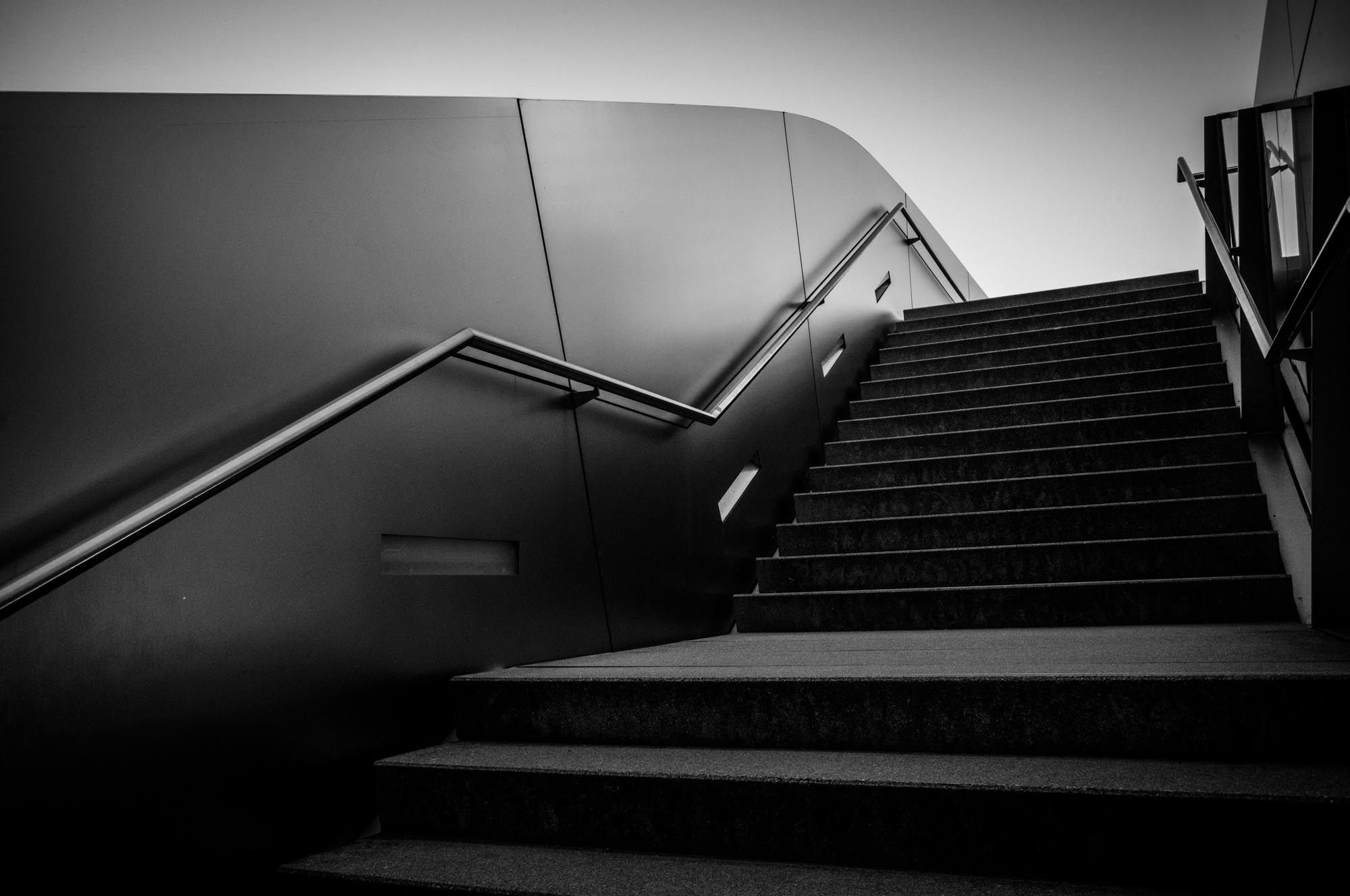 Black and white photo of a modern staircase and sleek railing in an urban setting, emphasizing architectural design.