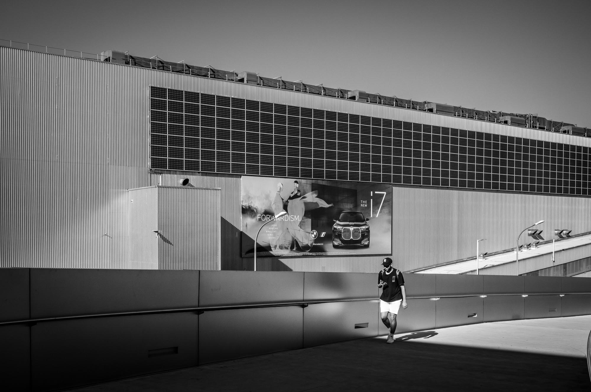 Person walking by a large building with a car advertisement, checking phone, urban scene in black and white.