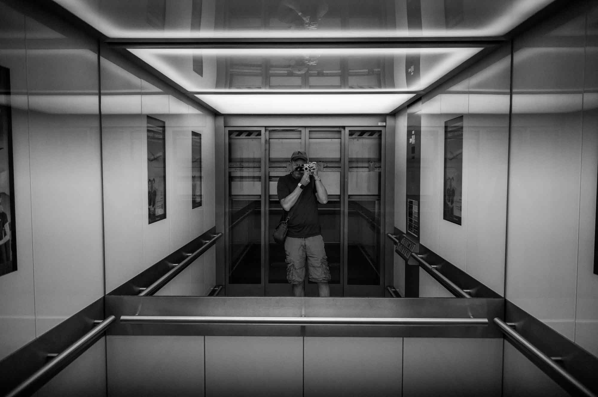Person taking a photo in a mirrored elevator interior, black and white.