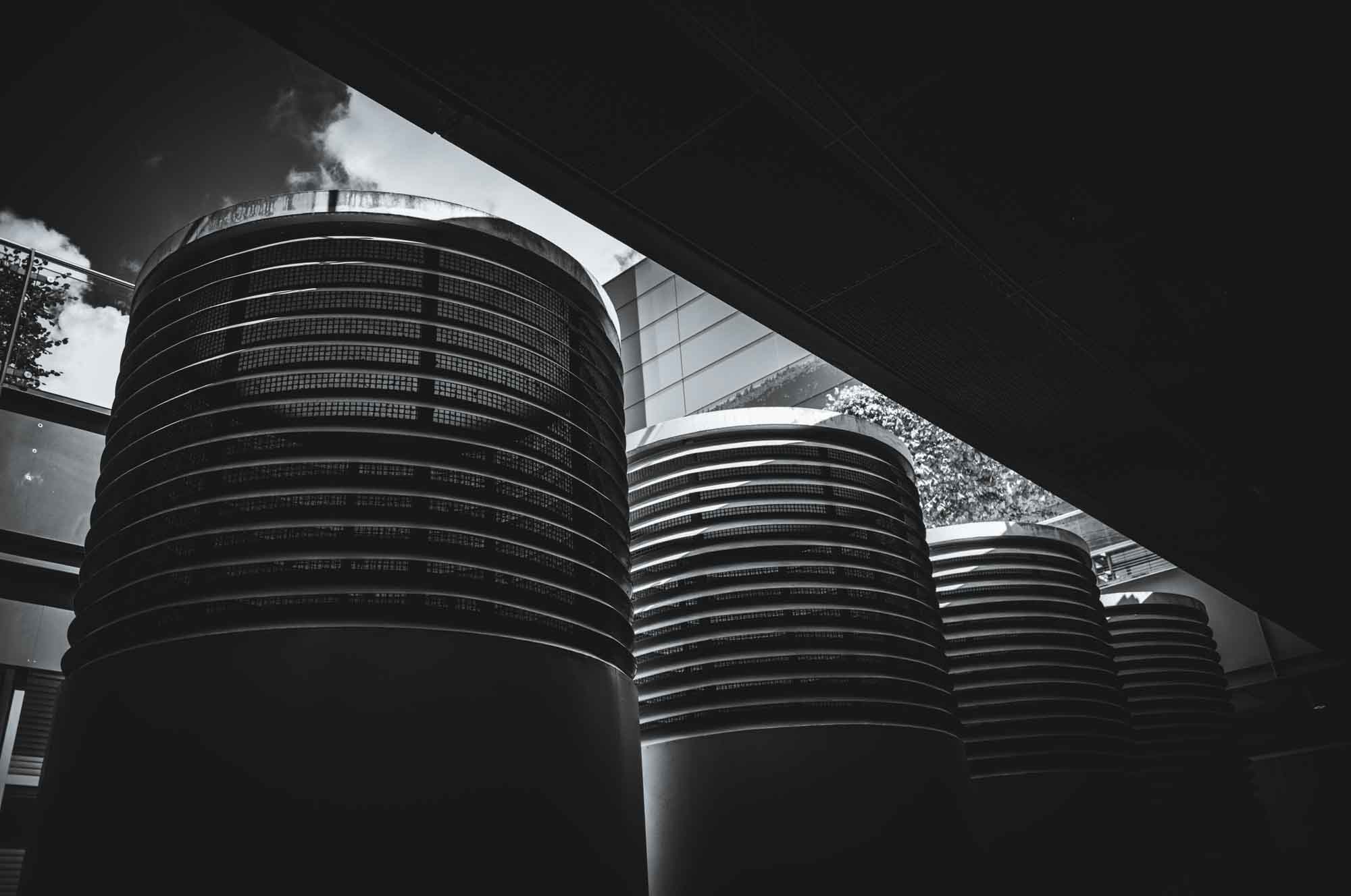 Black and white image of industrial cooling towers beneath a modern architectural structure with a cloudy sky backdrop.