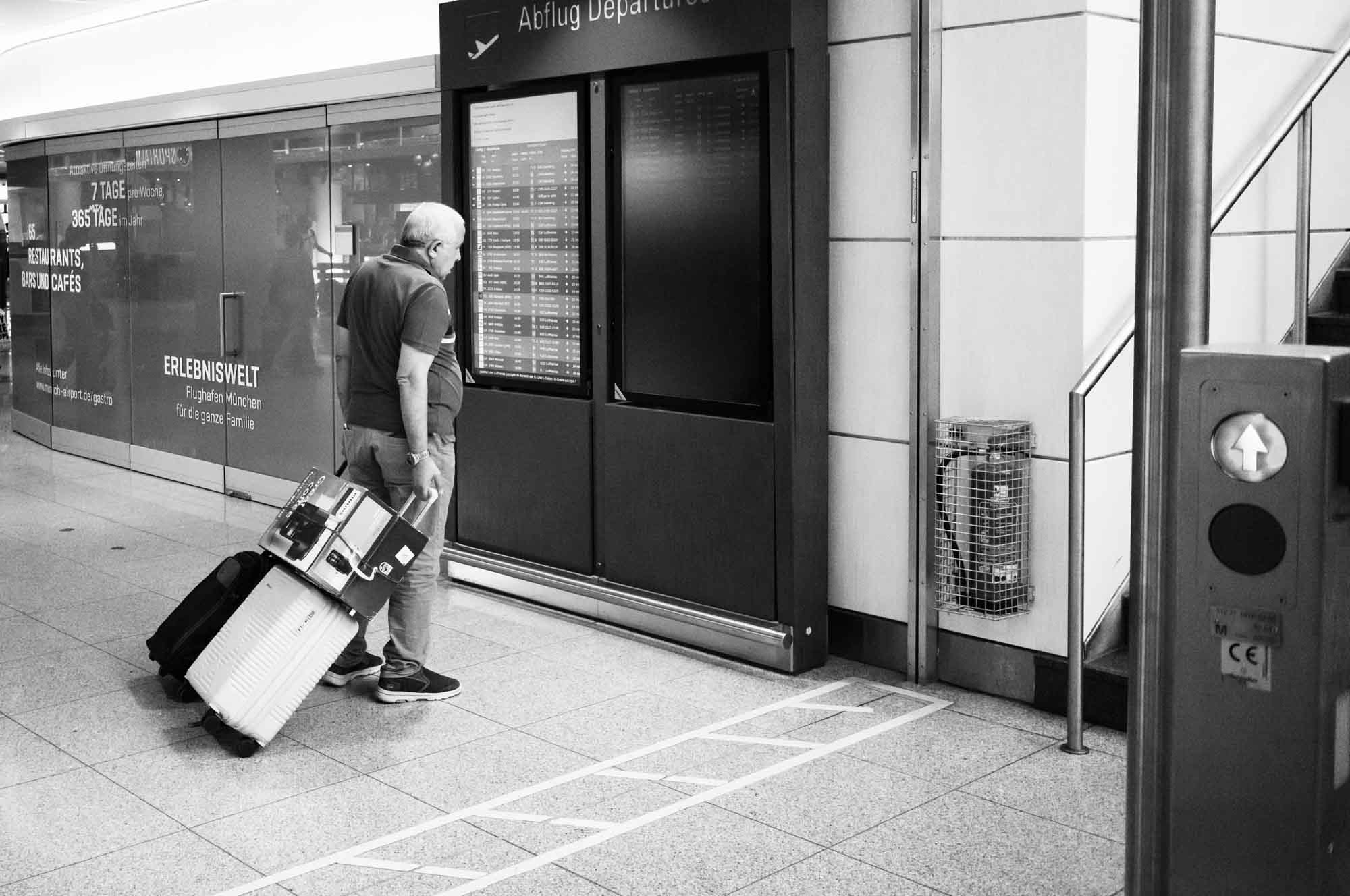 Traveler checks departure board at Munich Airport, pulling luggage.