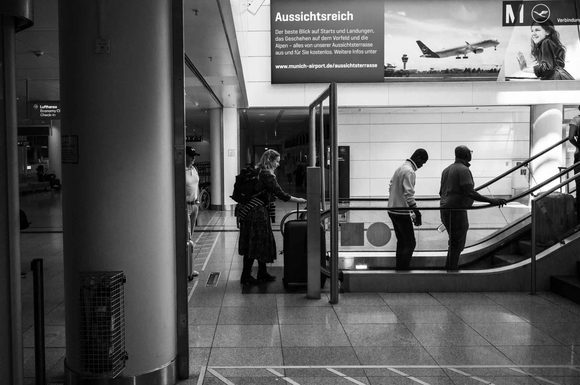 Passengers with luggage at escalator in Munich Airport terminal, black and white photo.