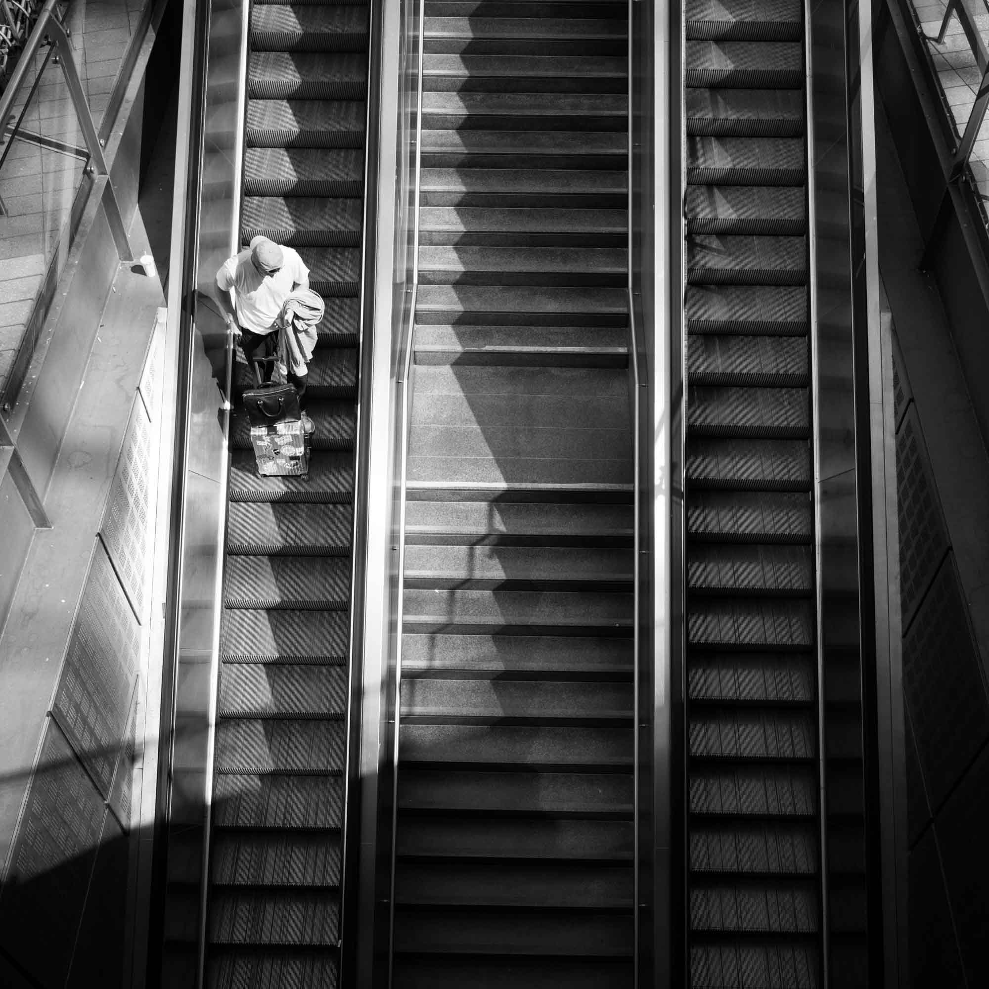 Person on the left escalator with luggage, black and white photo, high-angle view, urban scene shadows.