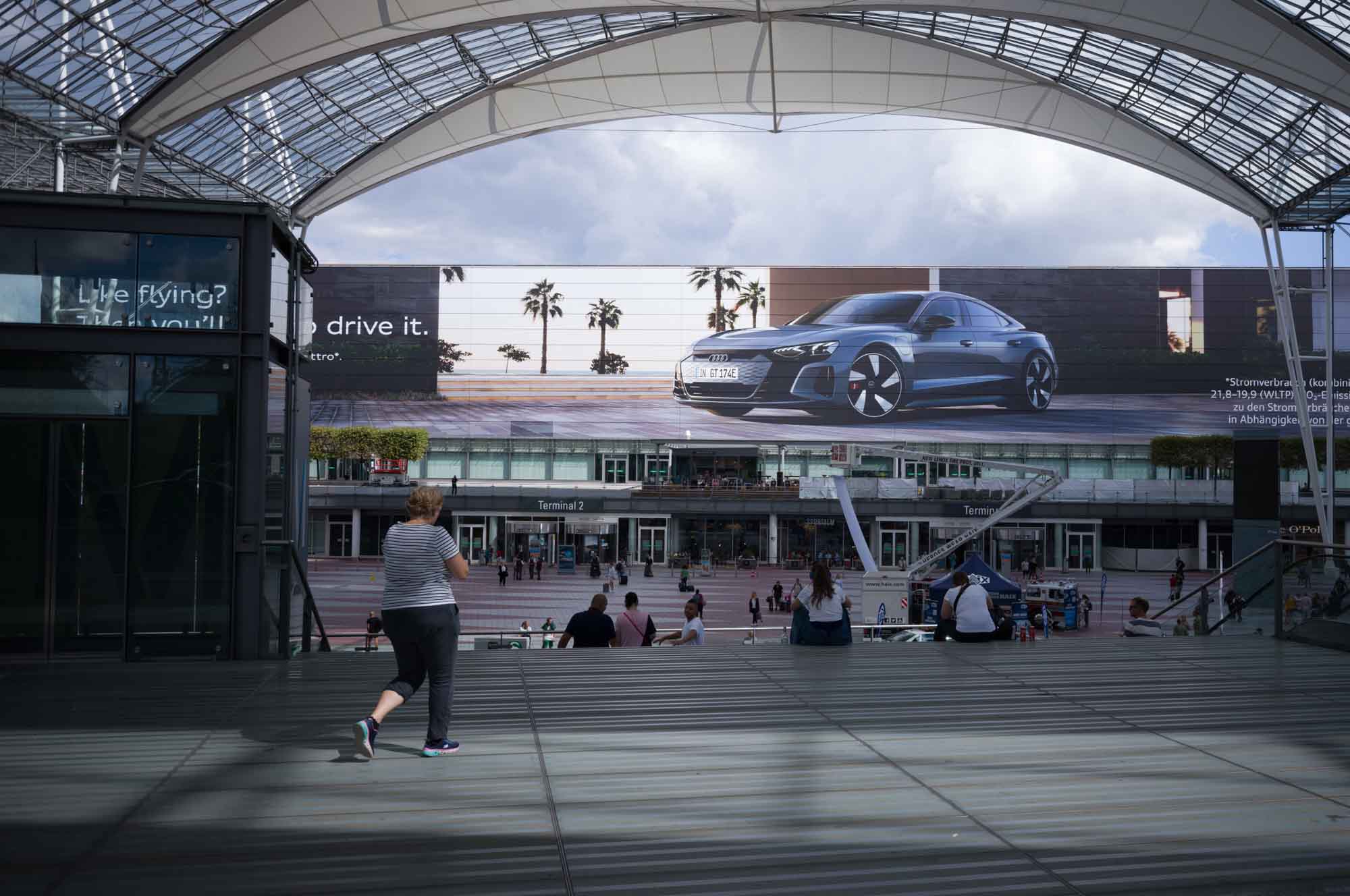 People walking under a large airport terminal canopy with a huge car advertisement in the background.