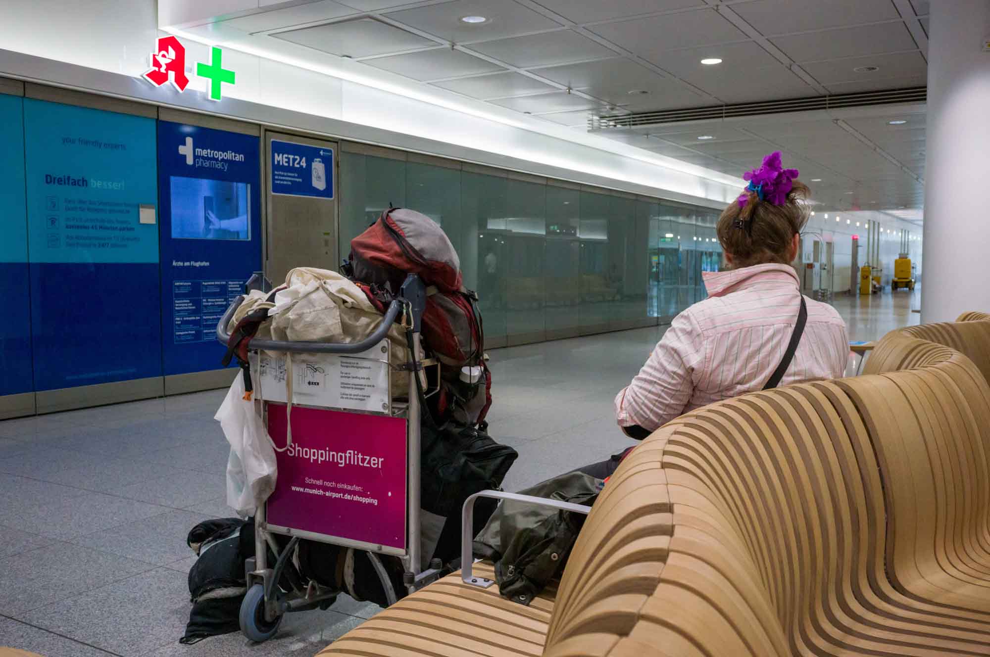 Traveler with packed trolley resting in an airport seating area near a pharmacy.