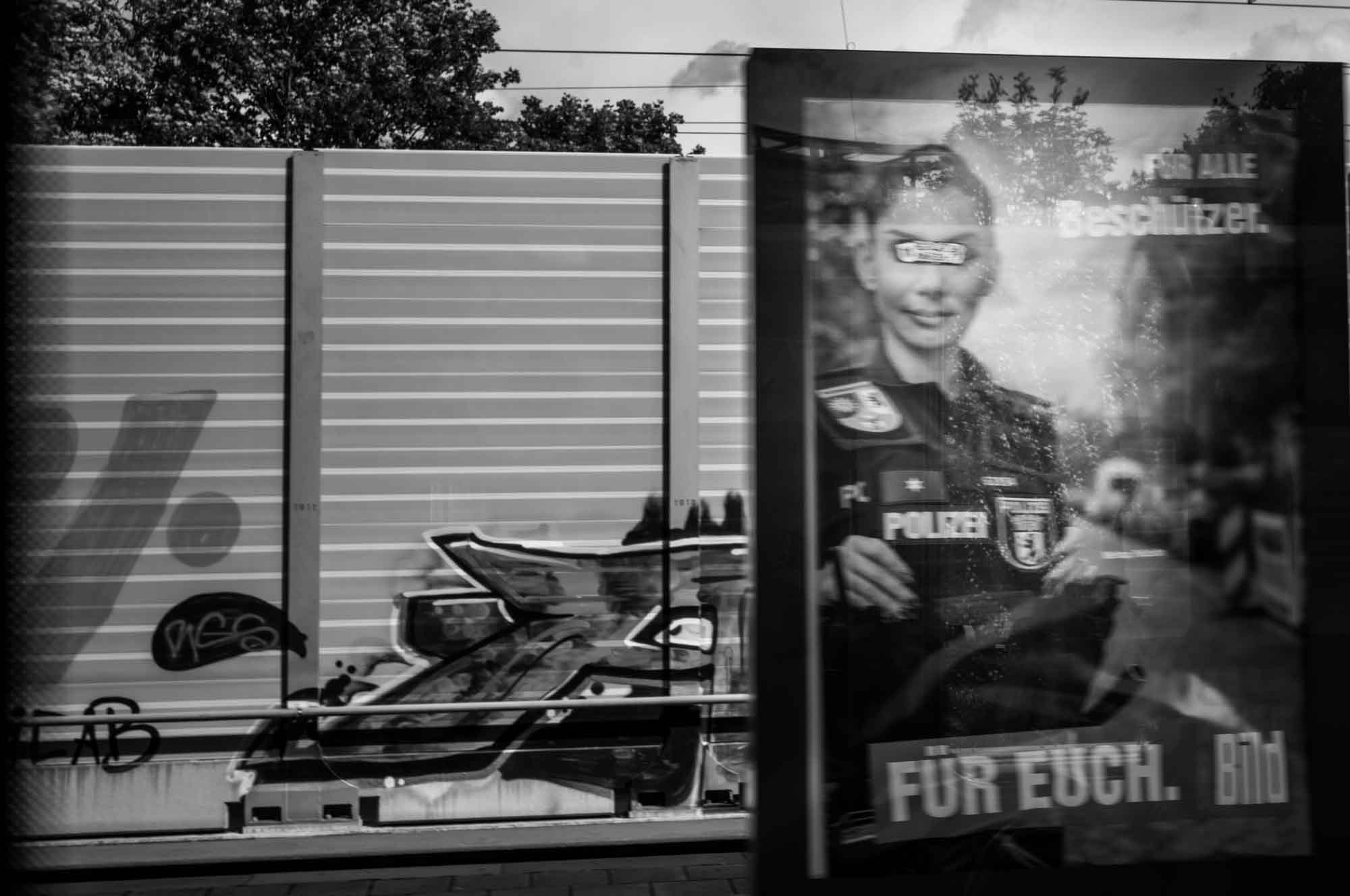 Black and white photo of graffiti-covered wall and a blurred police poster with reflective surface in urban setting.
