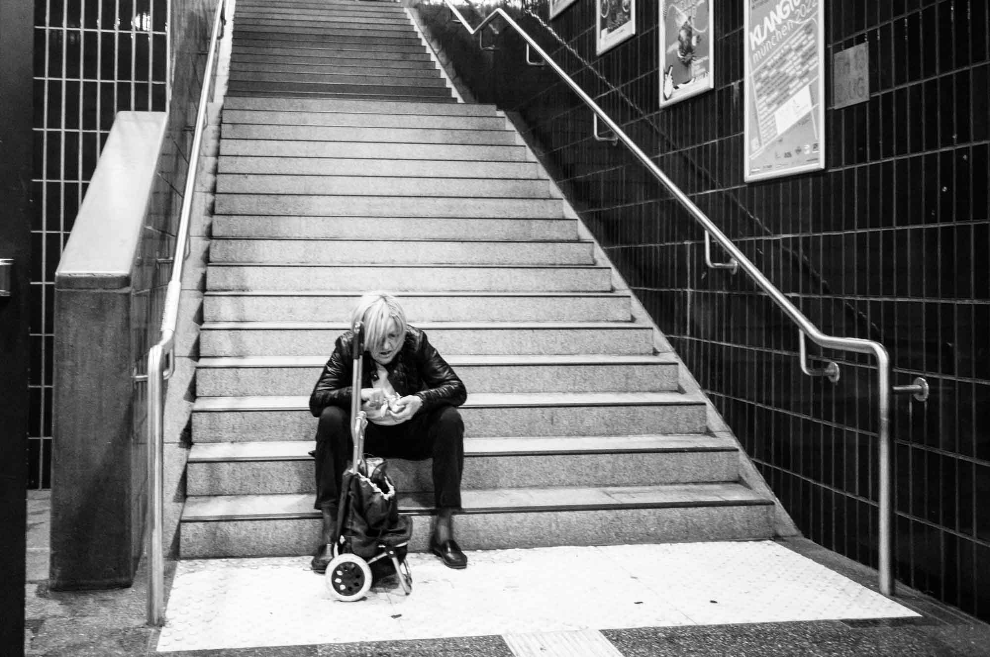 Person sitting on subway stairs with a shopping trolley, absorbed in thought, in a black and white urban setting.