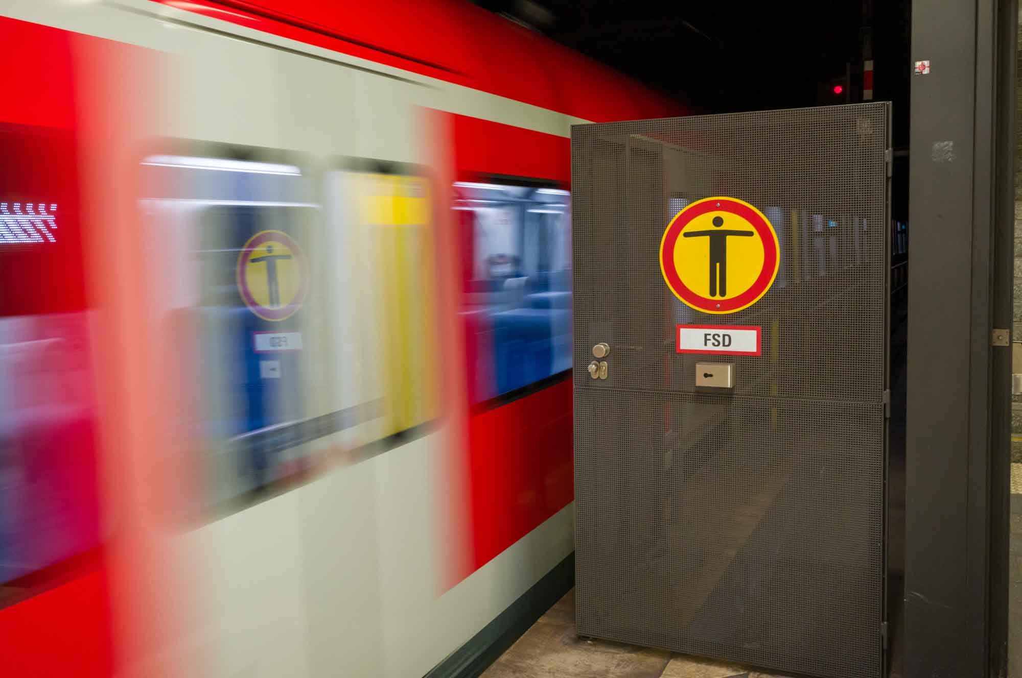Red train speeding past safety barrier with caution sign in a subway station.