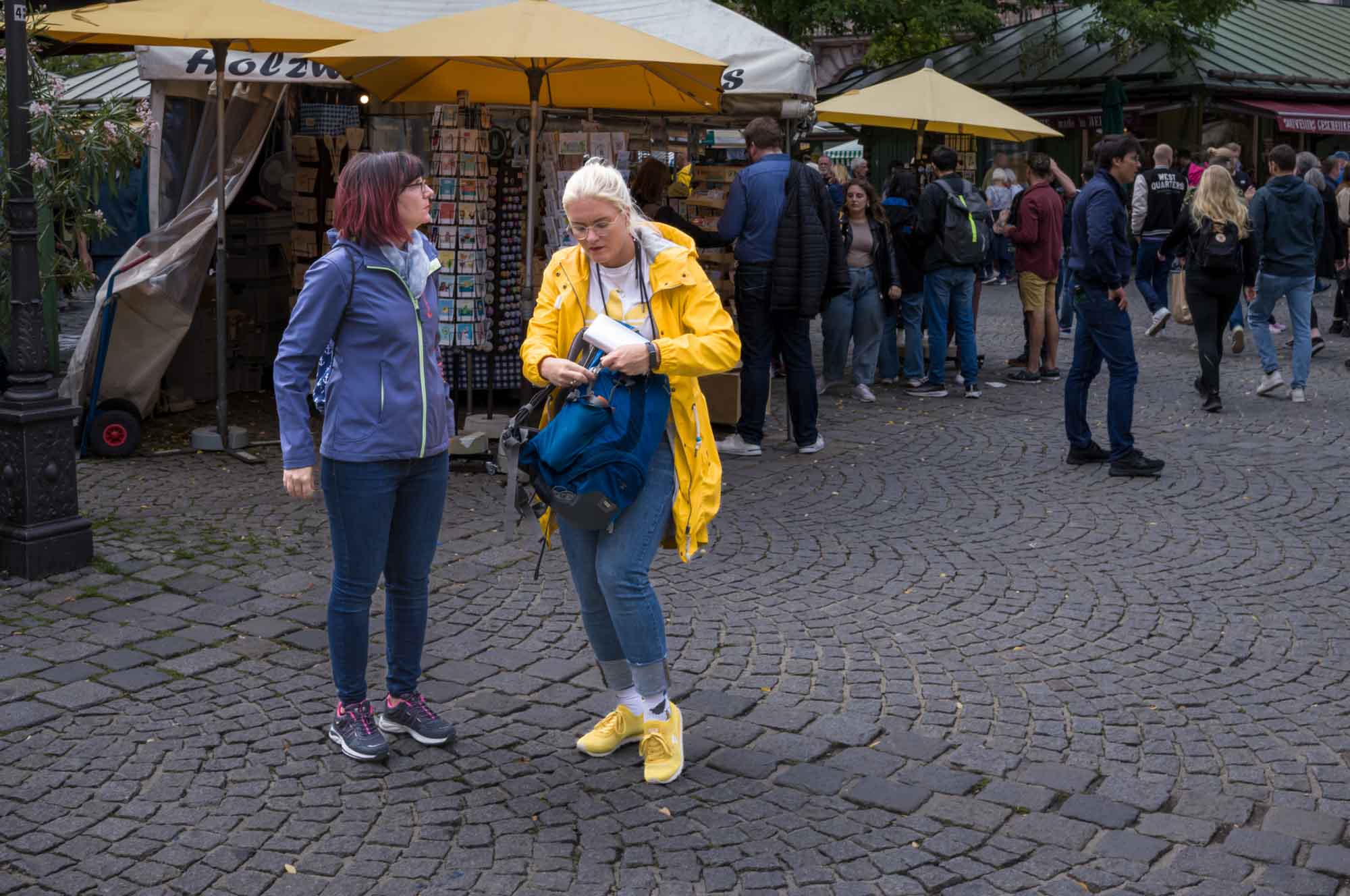 Two women stand on a cobblestone street, surrounded by market stalls and people, in casual outdoor wear.