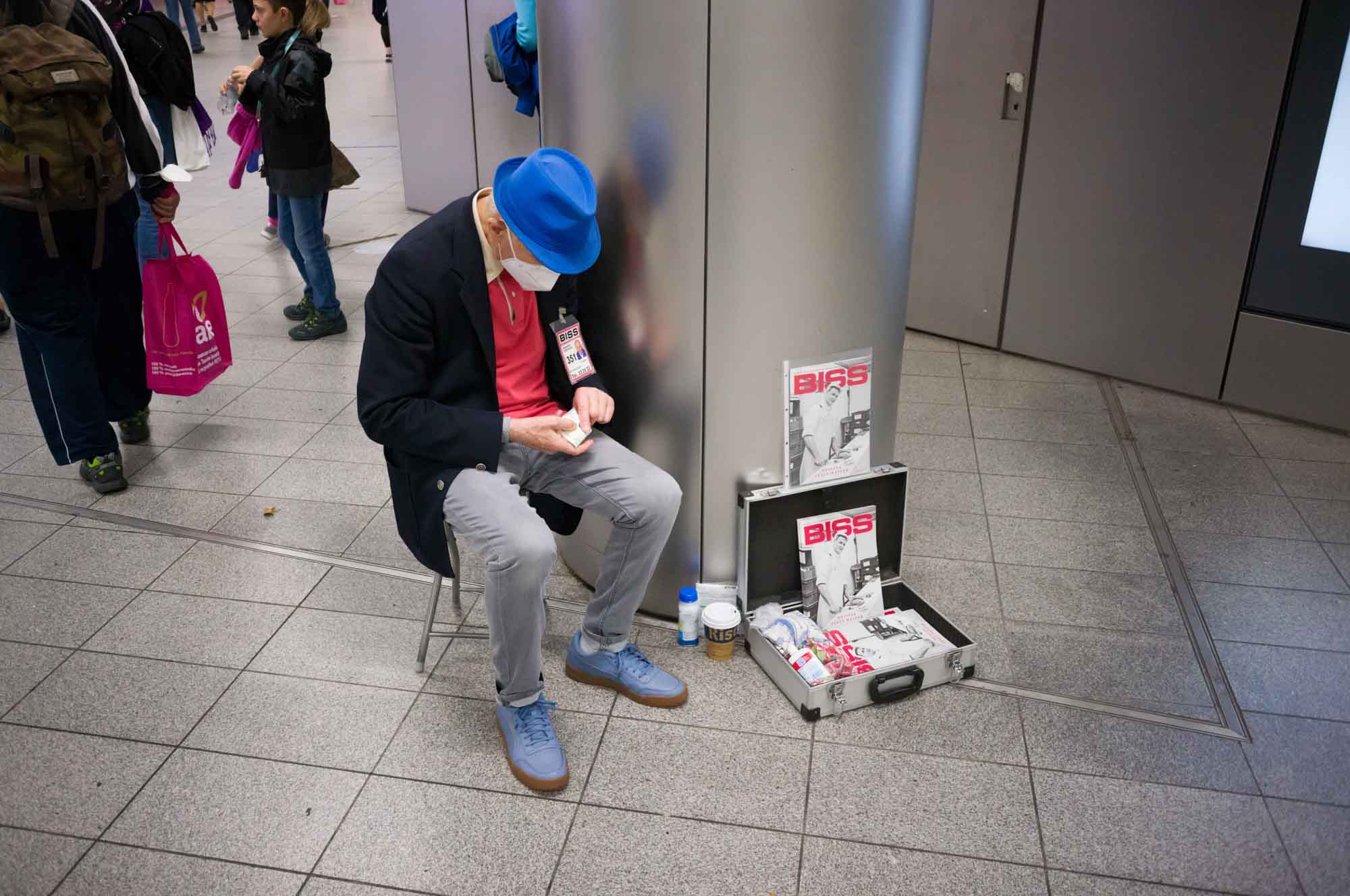 Man in blue hat selling magazines at train station; magazines displayed in open suitcase nearby.