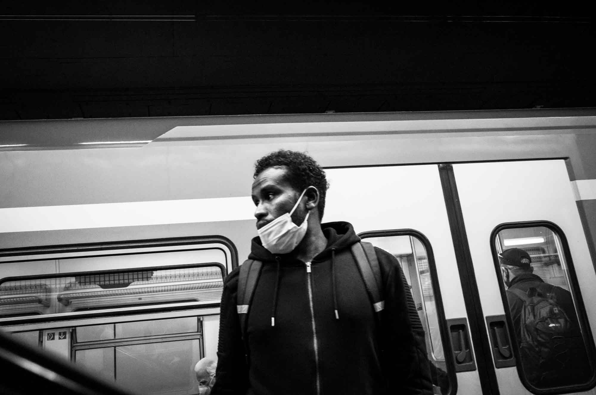 Man wearing a mask and backpack waits by a train in a station, black and white photography.
