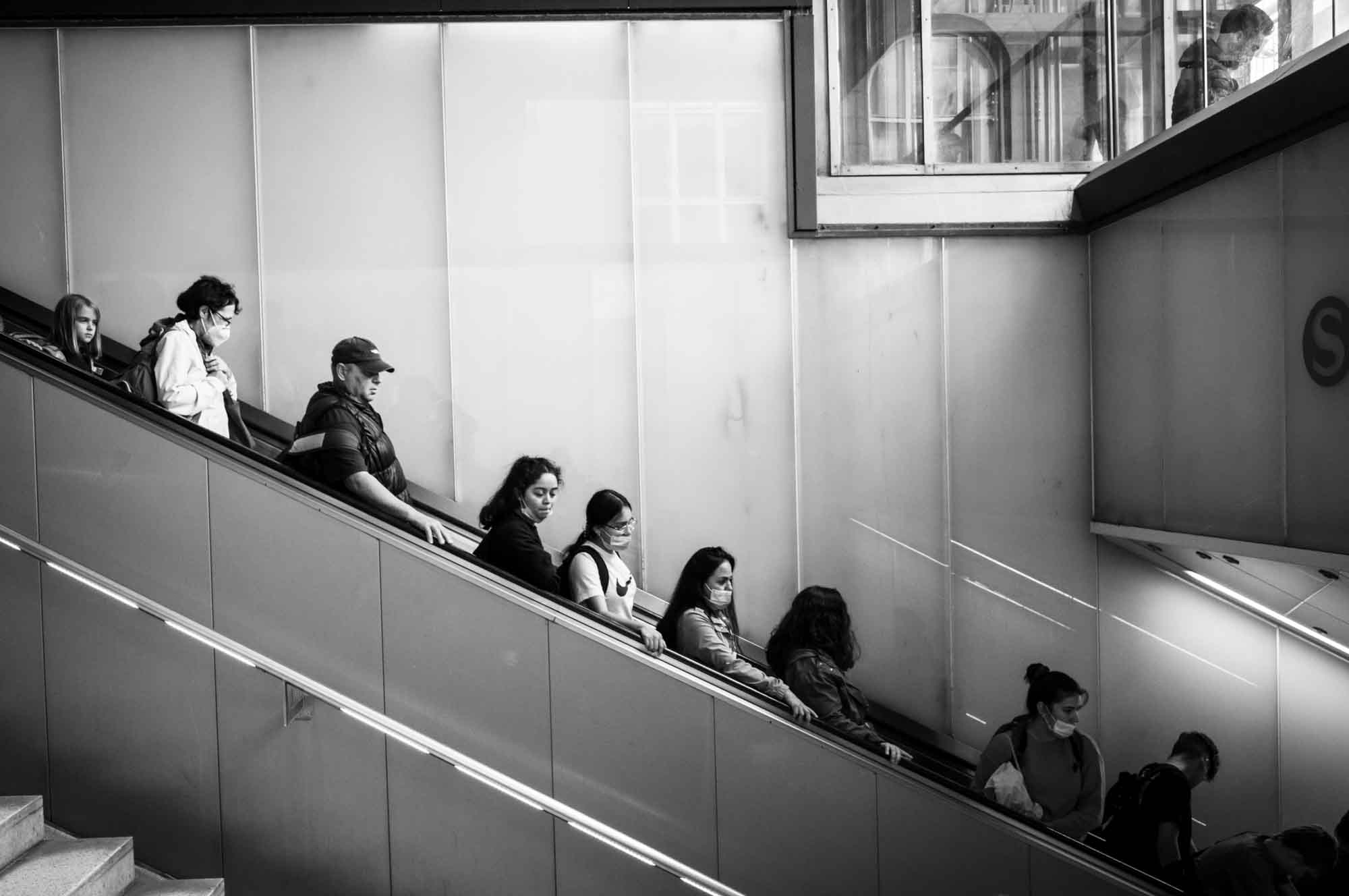 People descending on an indoor escalator, some wearing masks, in a modern building setting.