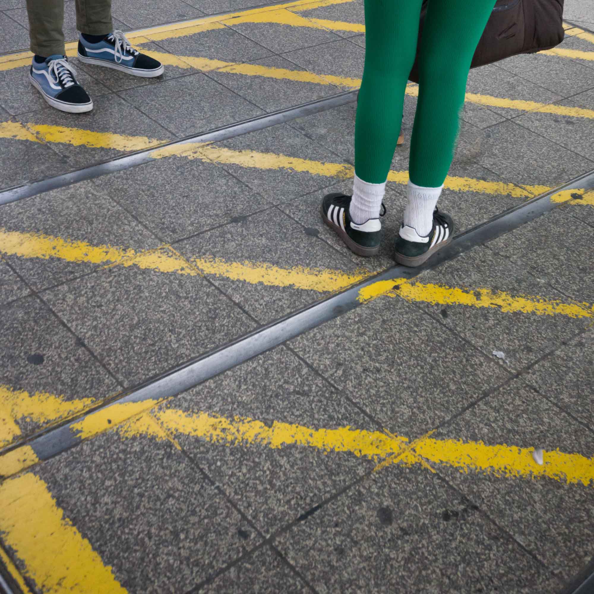 Two people standing on a platform with yellow safety lines. Casual footwear and green leggings visible.