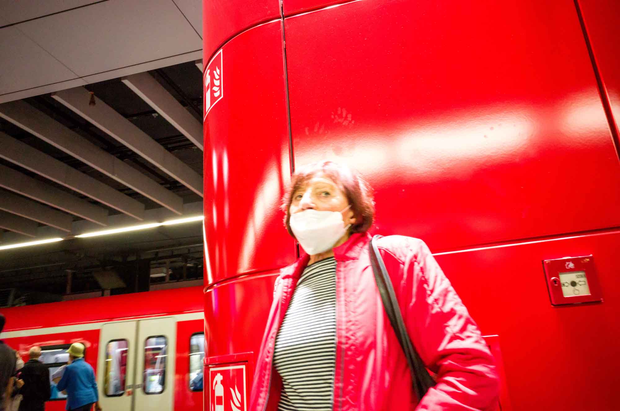 Person in a mask and red jacket at subway station with passing train in background.