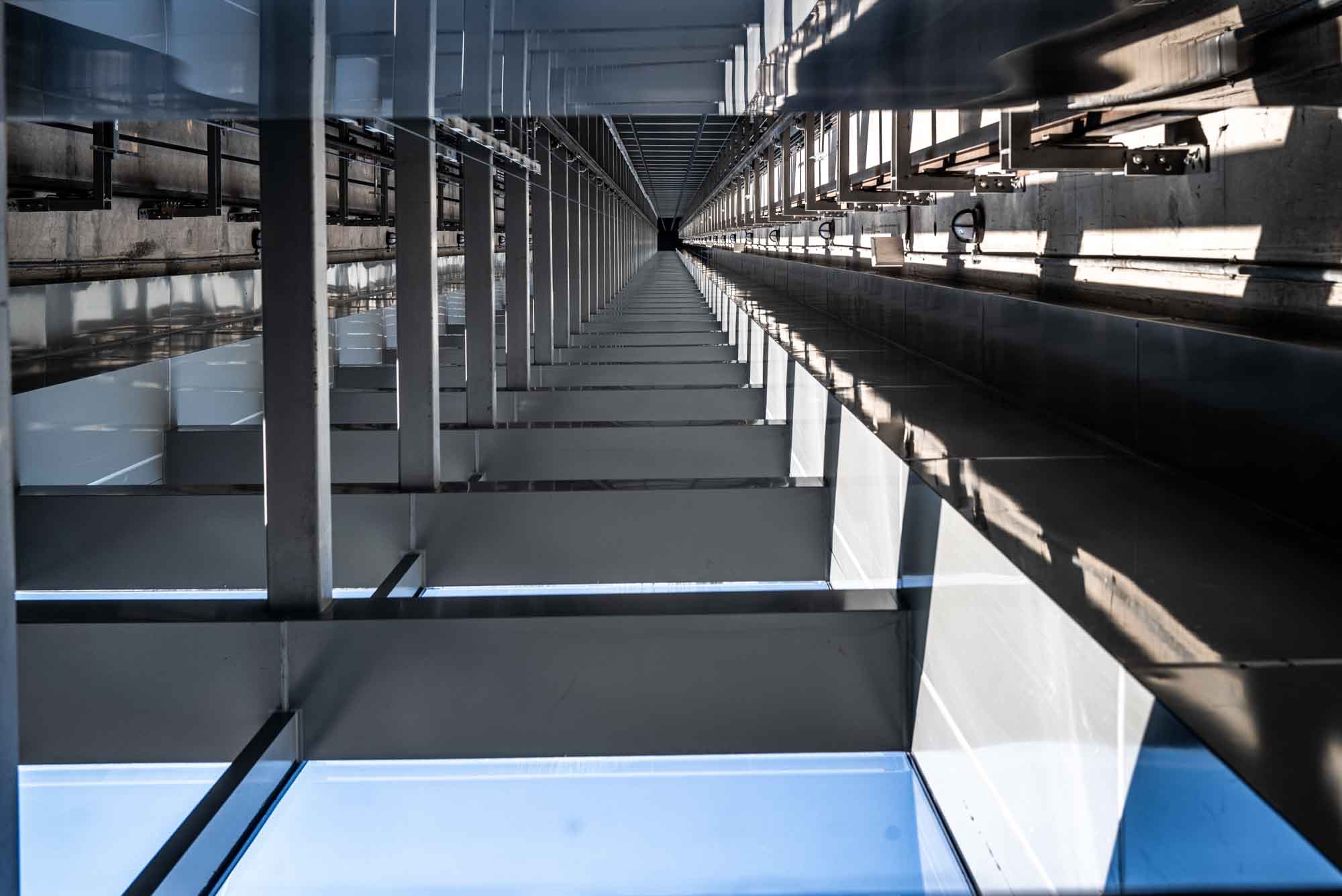 Looking up inside a tall, industrial elevator shaft with metal beams and reflected sunlight.