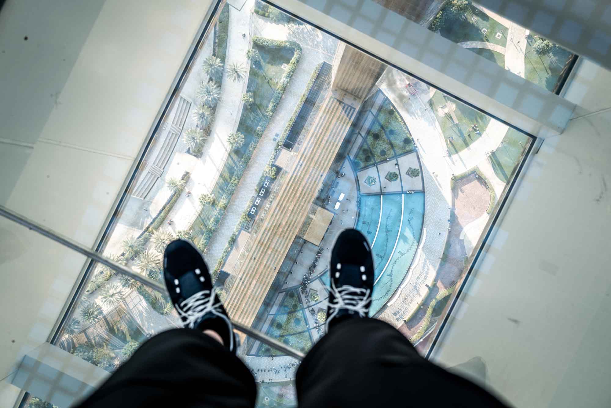 Looking down from a glass floor with a view of an outdoor plaza below, featuring a person’s feet in sneakers.
