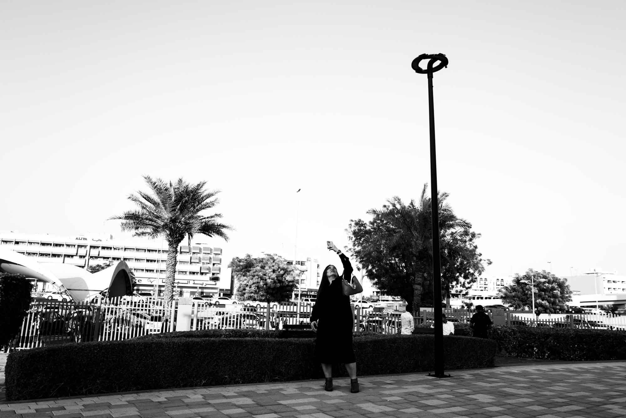 Person takes selfie against urban backdrop with palm trees and buildings in black and white setting.
