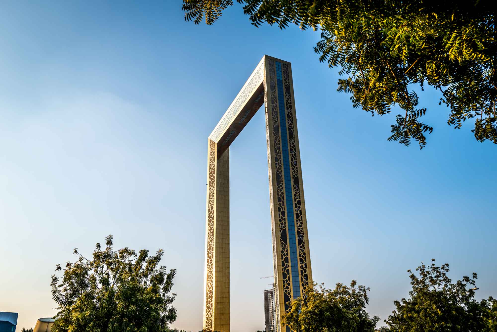 Iconic architectural landmark, Dubai Frame, against a clear blue sky, framed by lush trees.