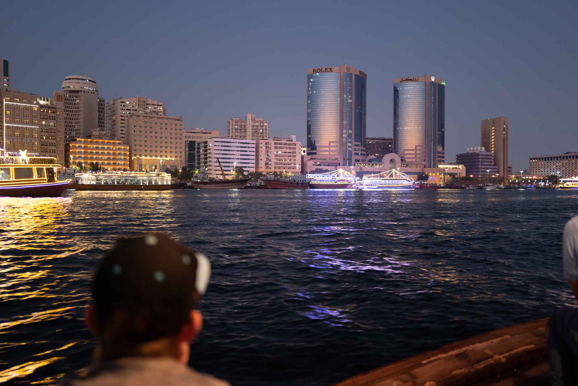 Dubai Creek at dusk with illuminated buildings and boats, viewed from a boat with a person in the foreground.