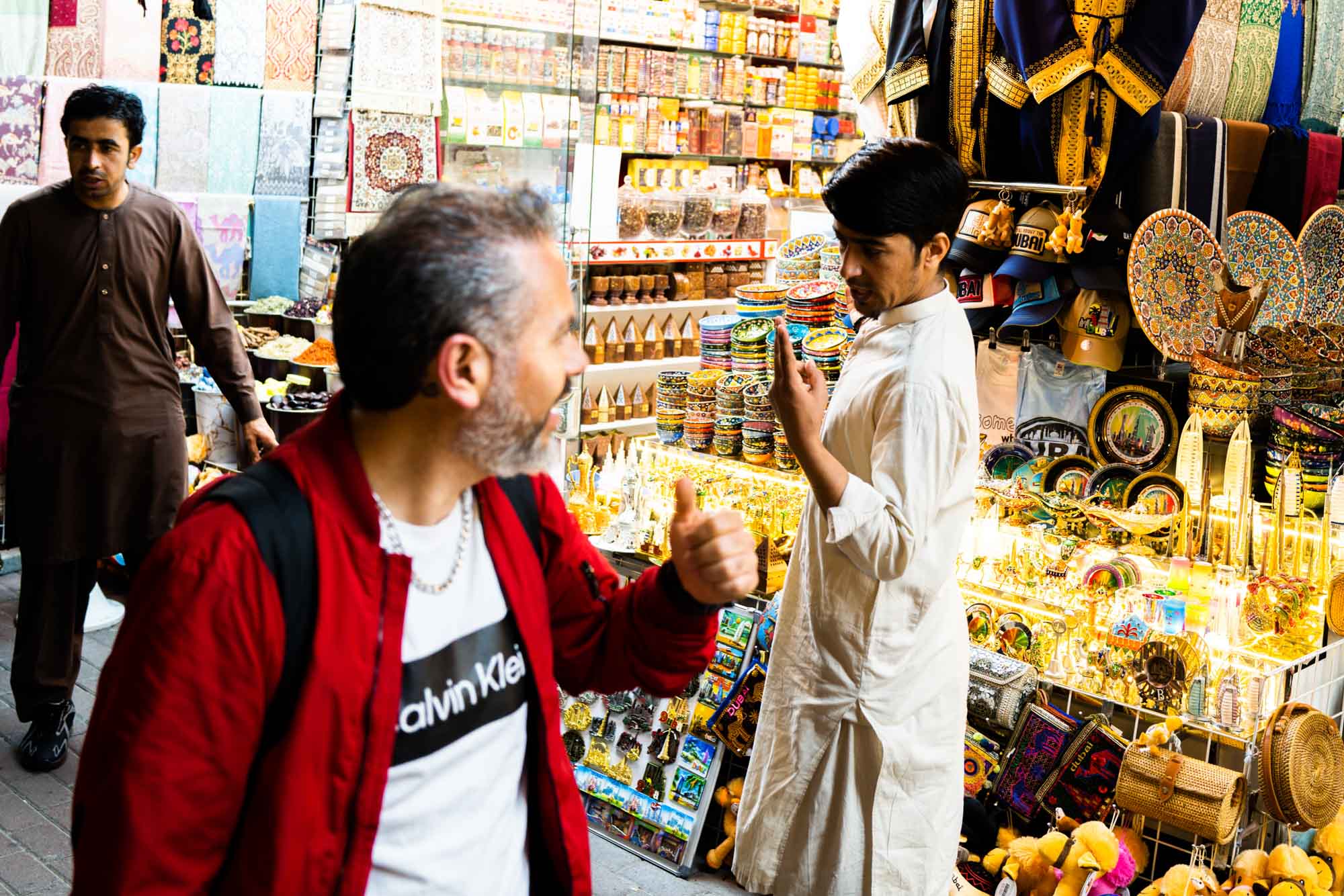 Man giving thumbs up in vibrant spice and souvenir market, colorful displays of crafts and textiles in background.