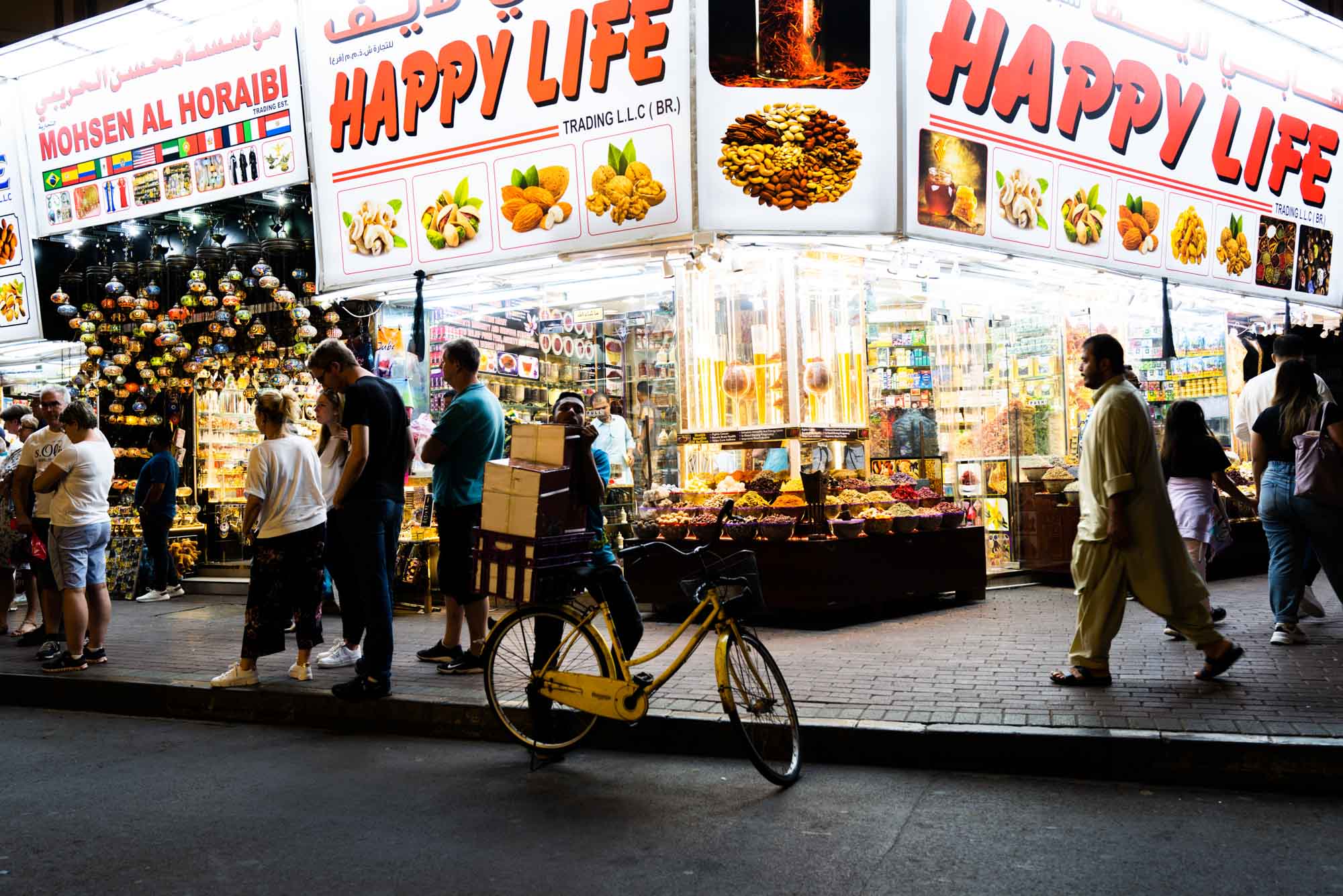 Busy market street scene with people shopping, a bicycle, and colorful lights at a vibrant food store at night.