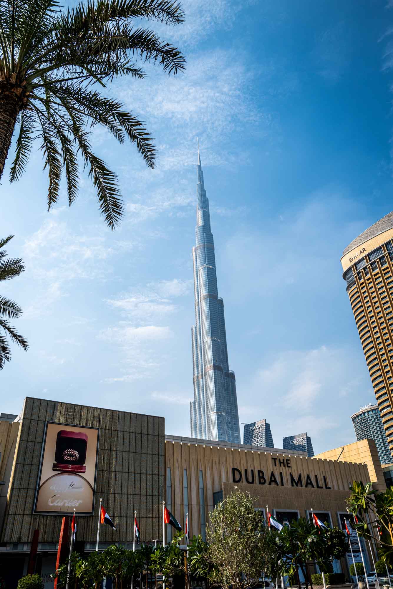 Dubai Mall with Burj Khalifa in the background on a sunny day, framed by palm trees and clear blue sky.