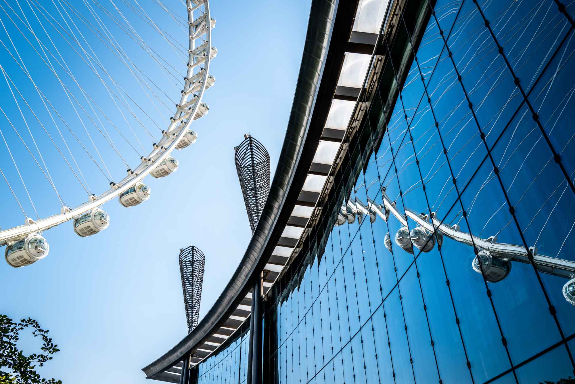 Modern ferris wheel and building reflection against blue sky, showcasing architectural contrast and design.