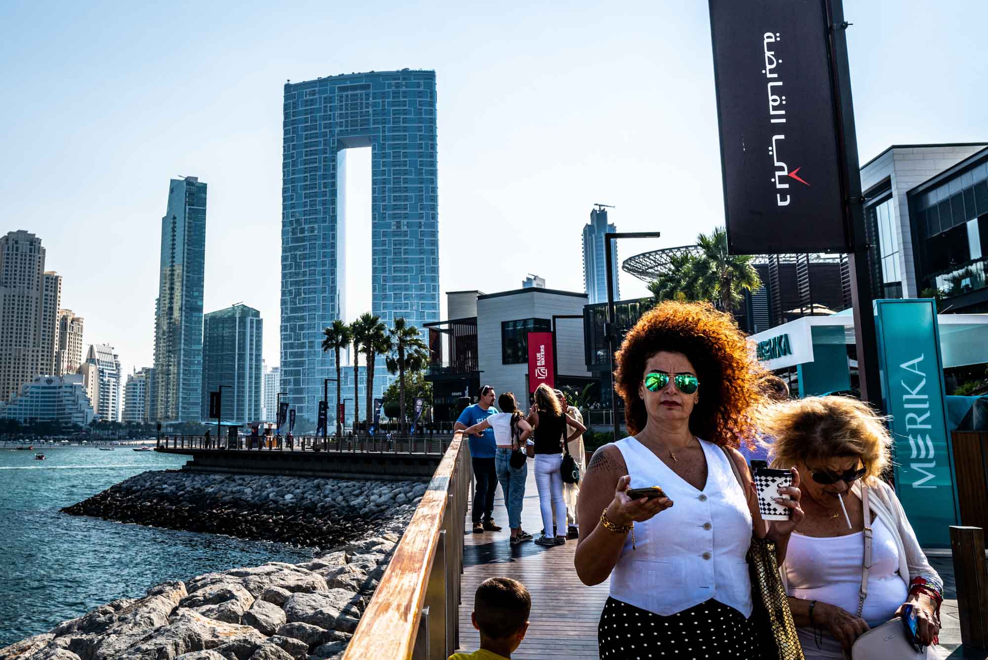 Modern Dubai skyline with people walking on a waterfront promenade, featuring unique high-rise architecture.