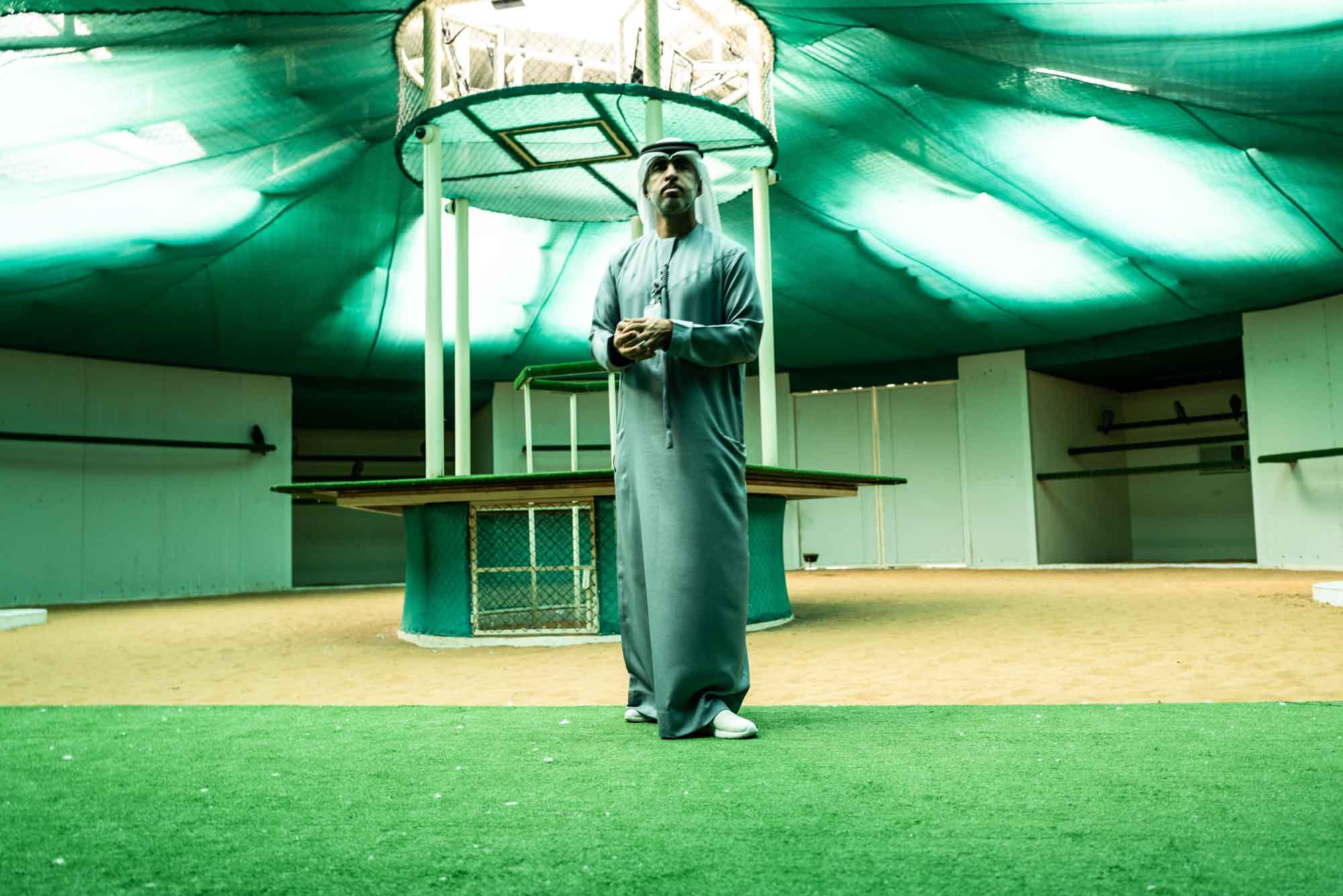 Man in traditional attire stands under a green canopy, surrounded by a sandy and grassy area.