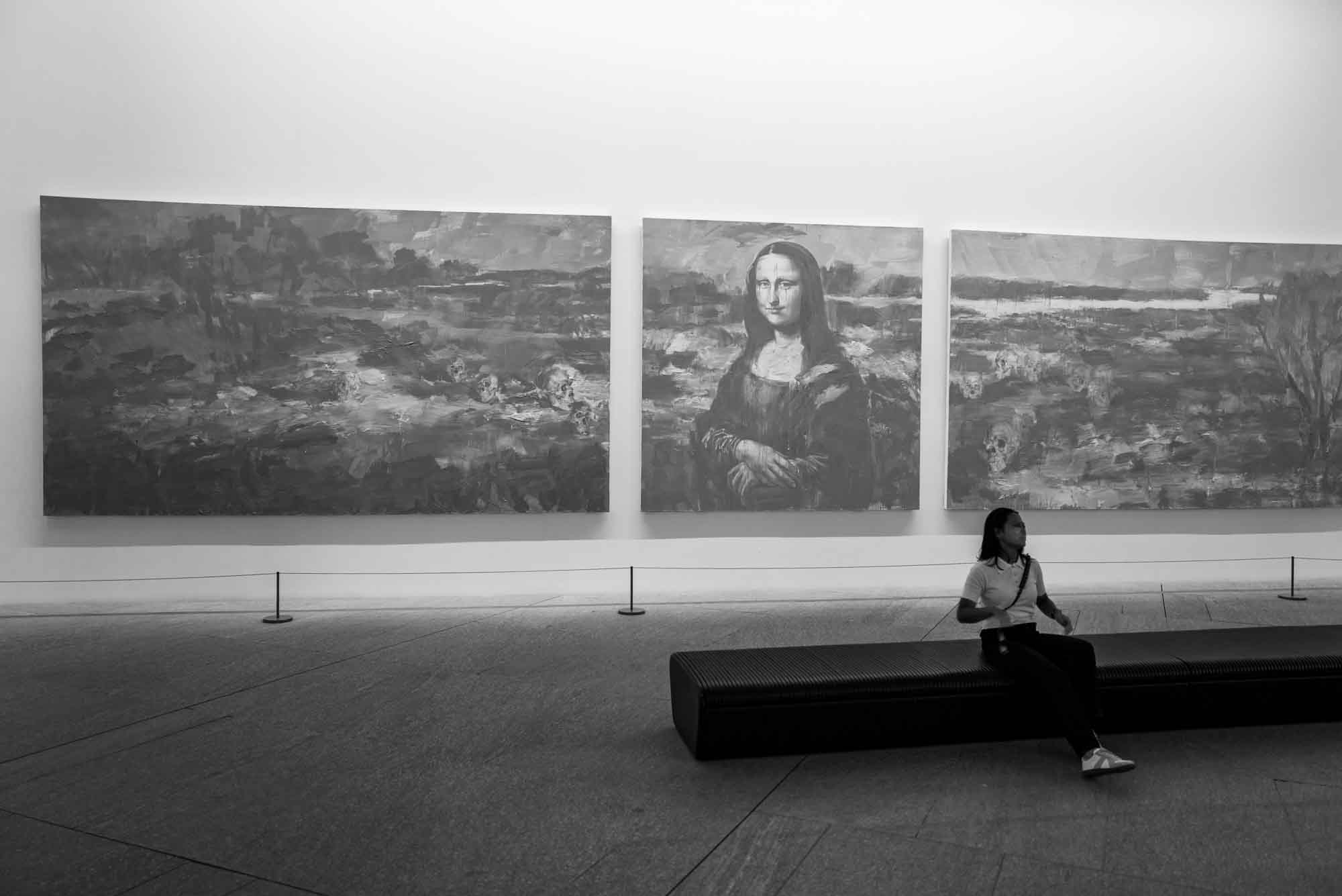 Person sitting on bench in art gallery, viewing a large triptych including Mona Lisa portrait. Black and white photograph.