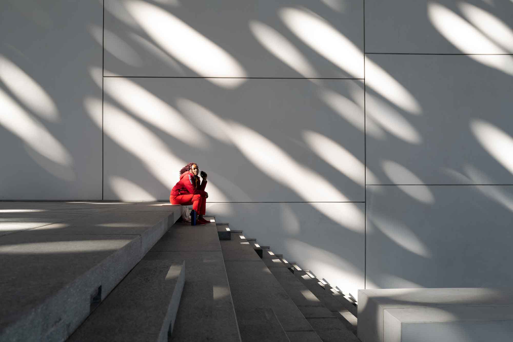 Person in red outfit sitting on concrete steps, surrounded by patterned shadows on a sunny day.