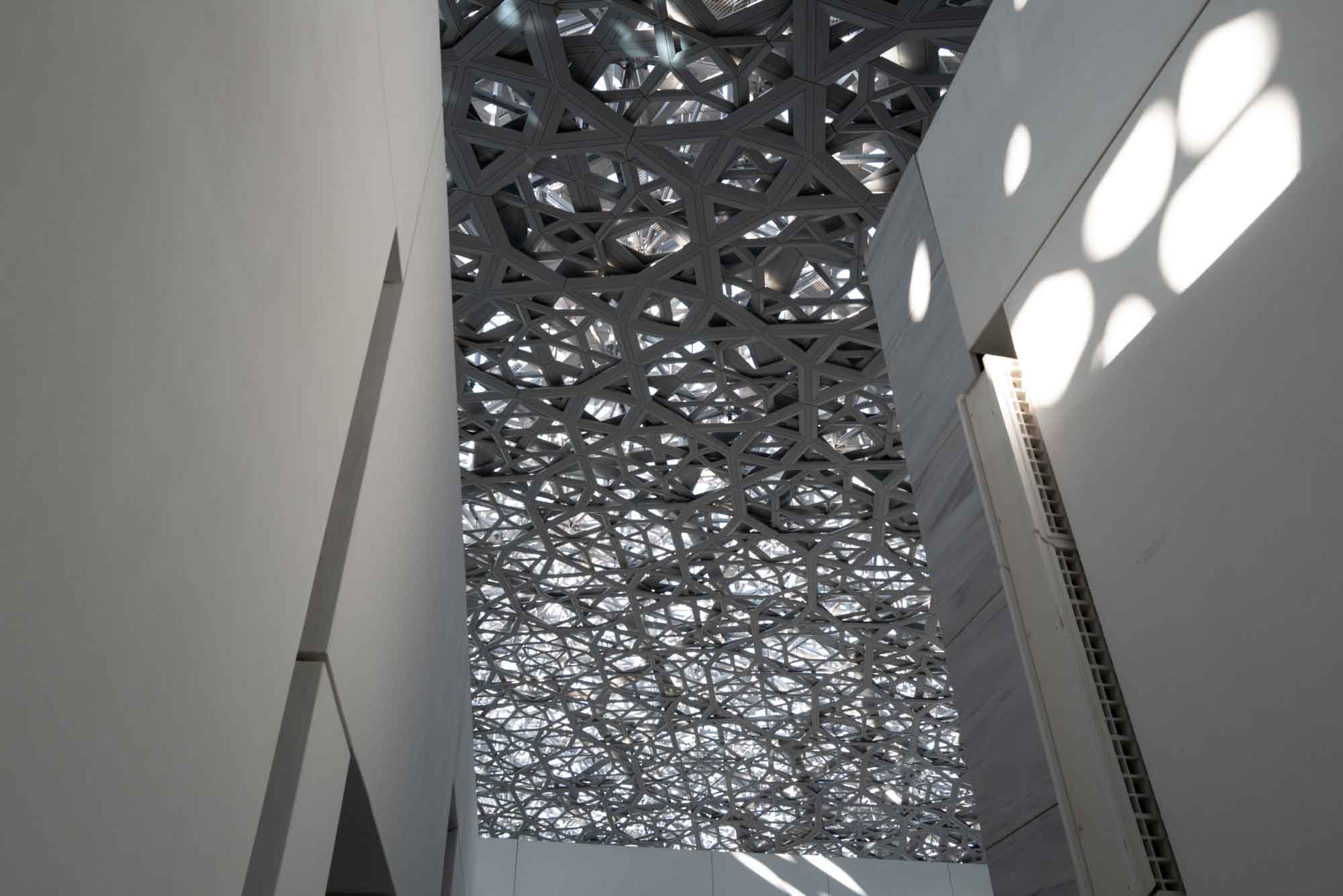 Intricate geometric ceiling design with sunlight casting shadows on white walls in a modern architectural space.