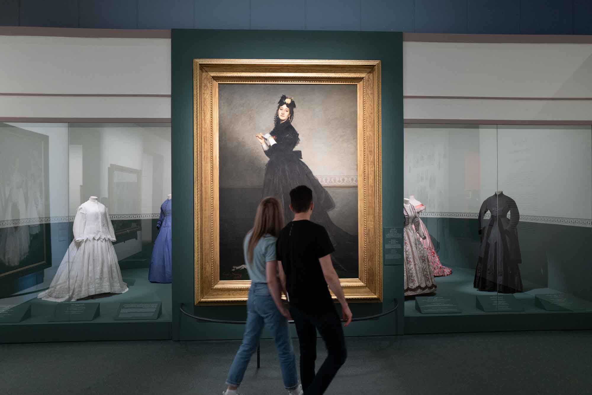 Visitors admire a large portrait in a museum exhibit featuring historical dresses on display.