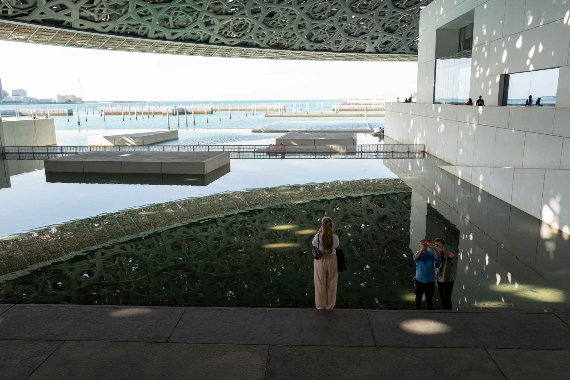 Modern architectural design overlooking waterfront with people enjoying the view under intricate patterned ceiling.