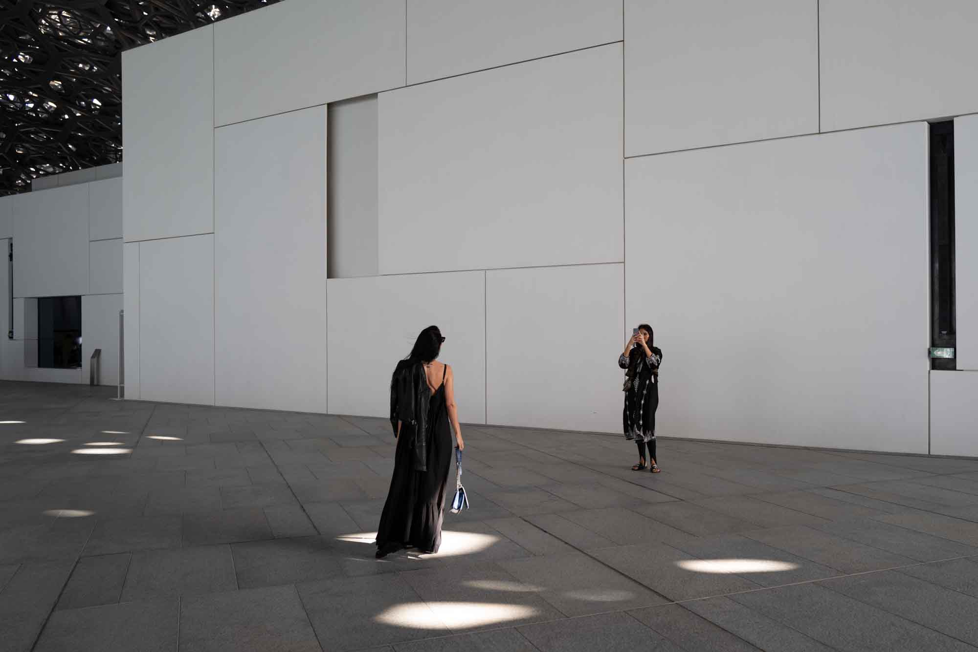 Two women in black outfits at a modern architectural space, one taking a photo of the other.