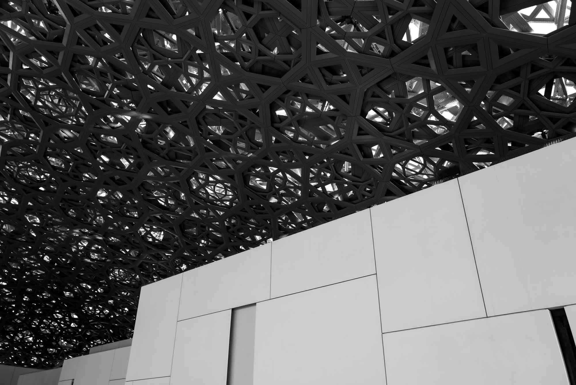 Intricate geometric ceiling above modern white building exterior, showcased in black and white.