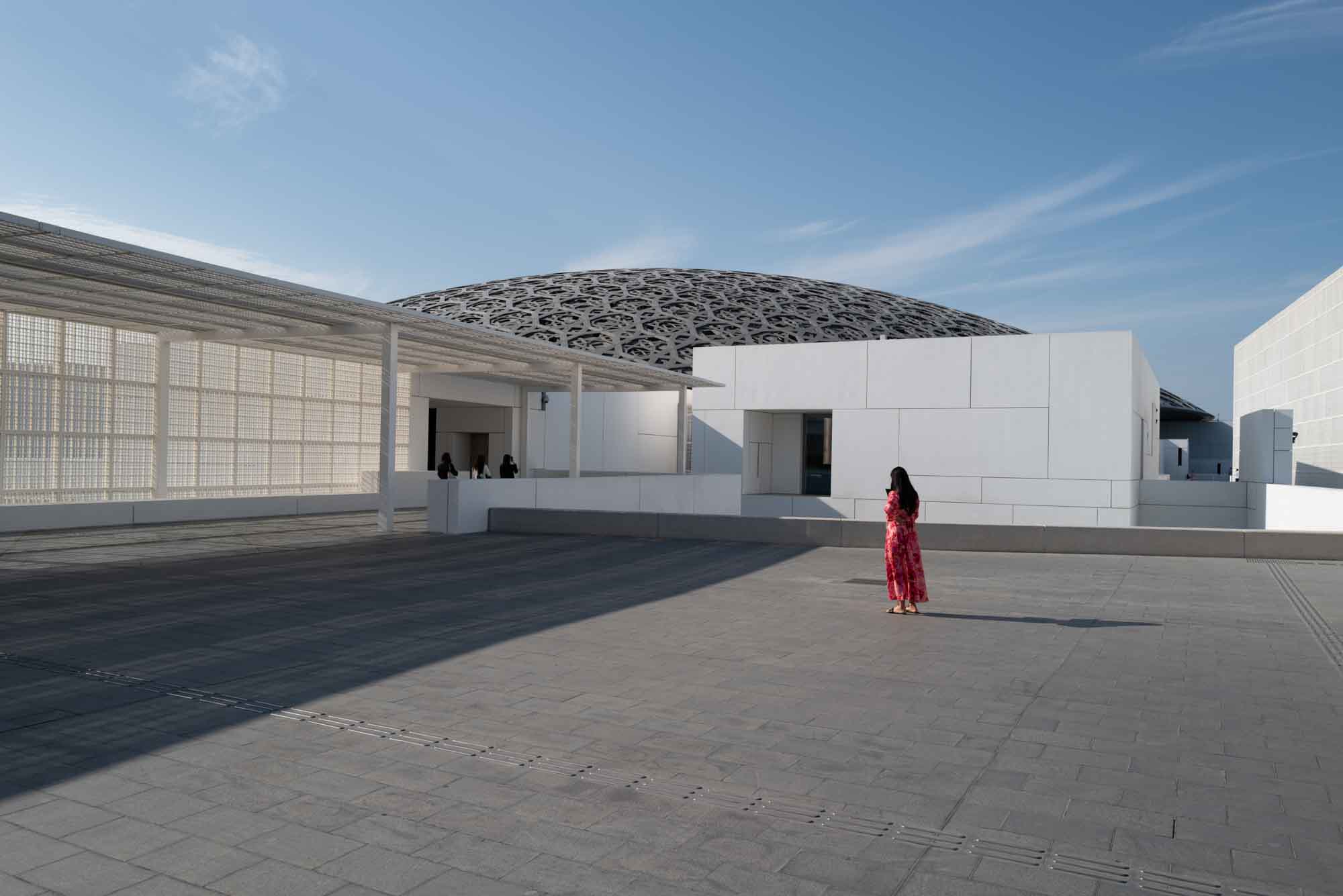 Woman in red dress explores modern architecture under clear blue sky.