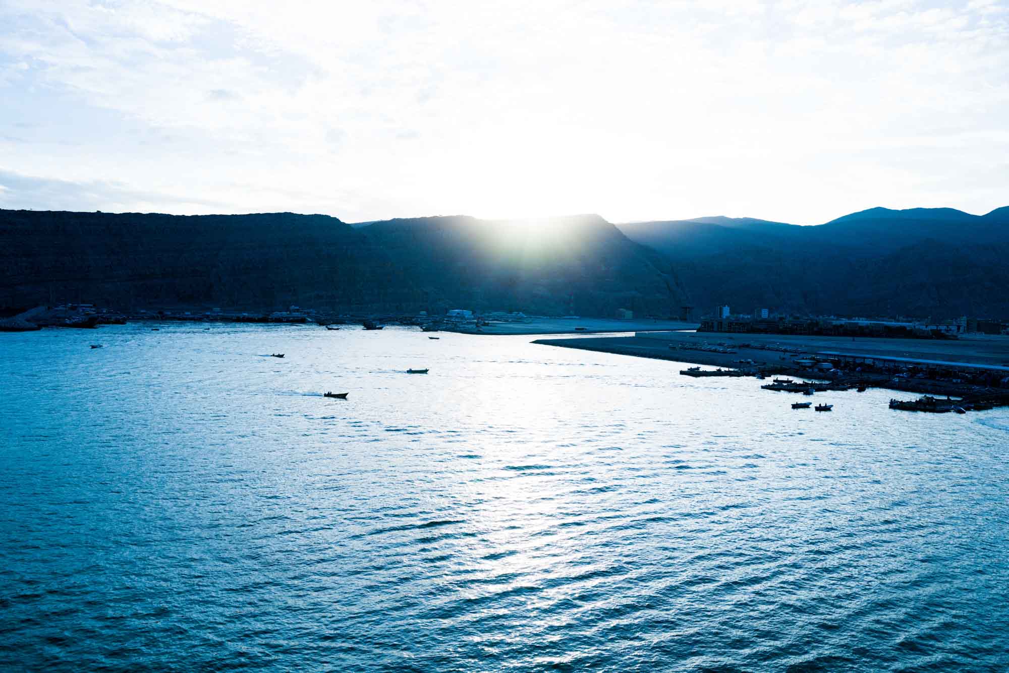 Sunrise over a coastal harbor with boats on calm waters, mountains silhouetted in the background.