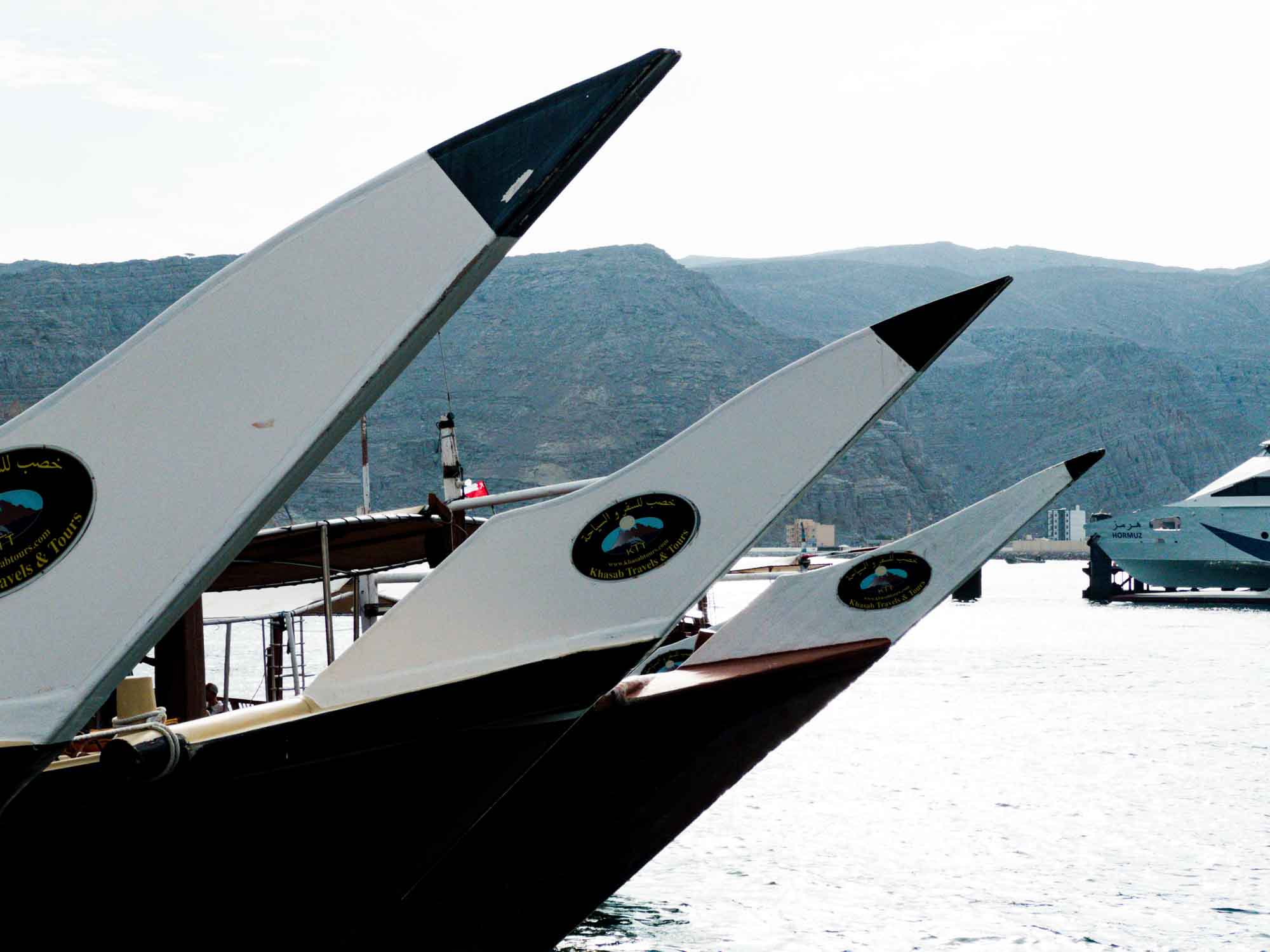 Traditional wooden dhow boats lined up in a scenic harbor with mountainous backdrop.