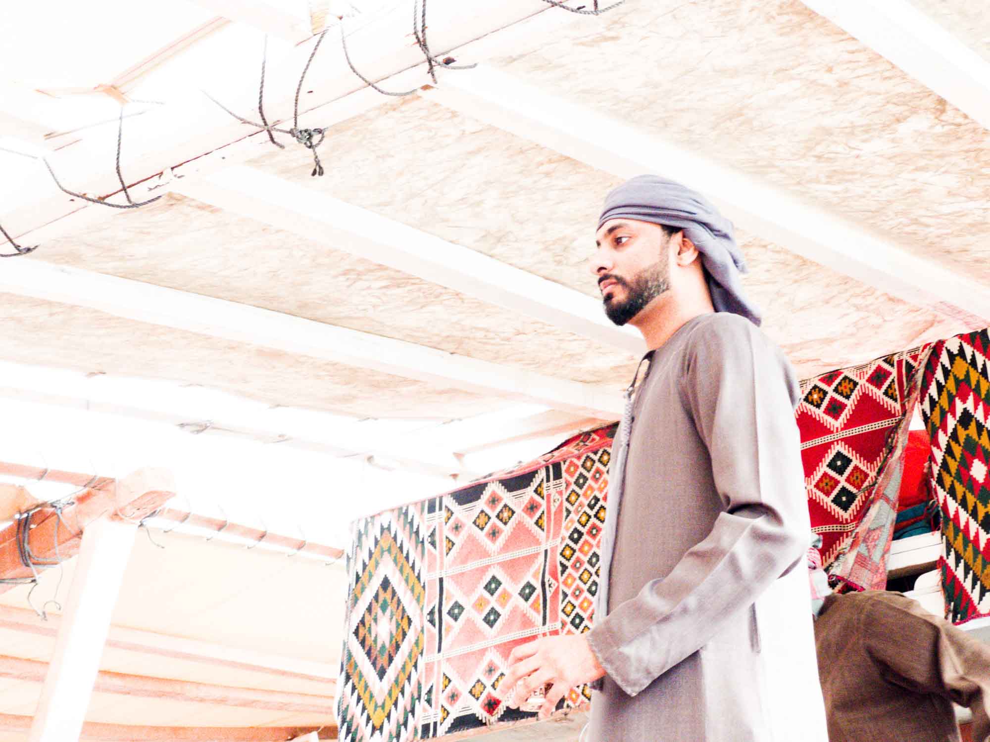Man in traditional attire with patterned textiles in the background, under wooden roof beams.