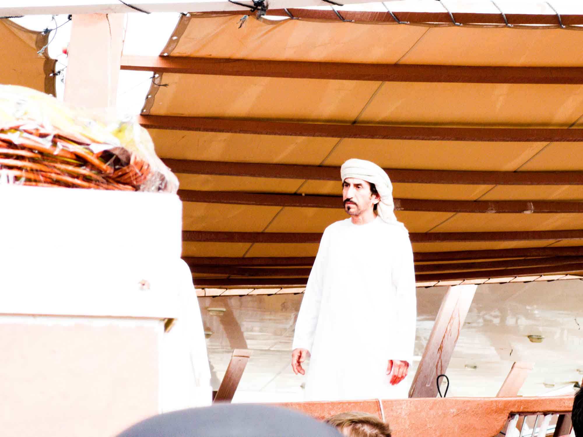 Man in traditional attire standing on a wooden platform under a canopy, with a basket in the foreground.